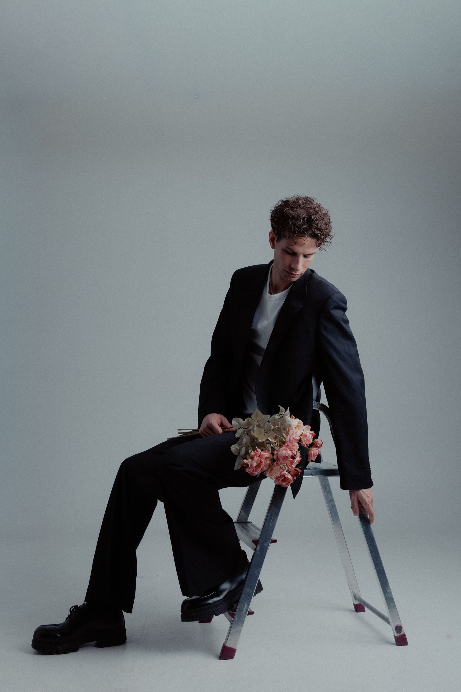 Stylish man in black suit sitting on a ladder holding wilted pink flowers on grey background.