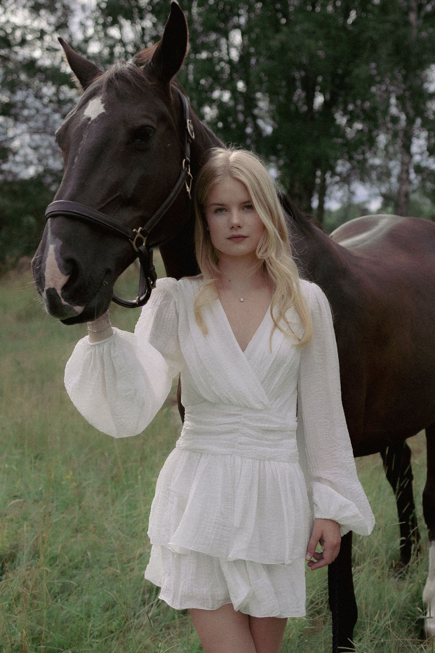 Young blonde woman in a white dress standing beside a dark horse in a rural field.