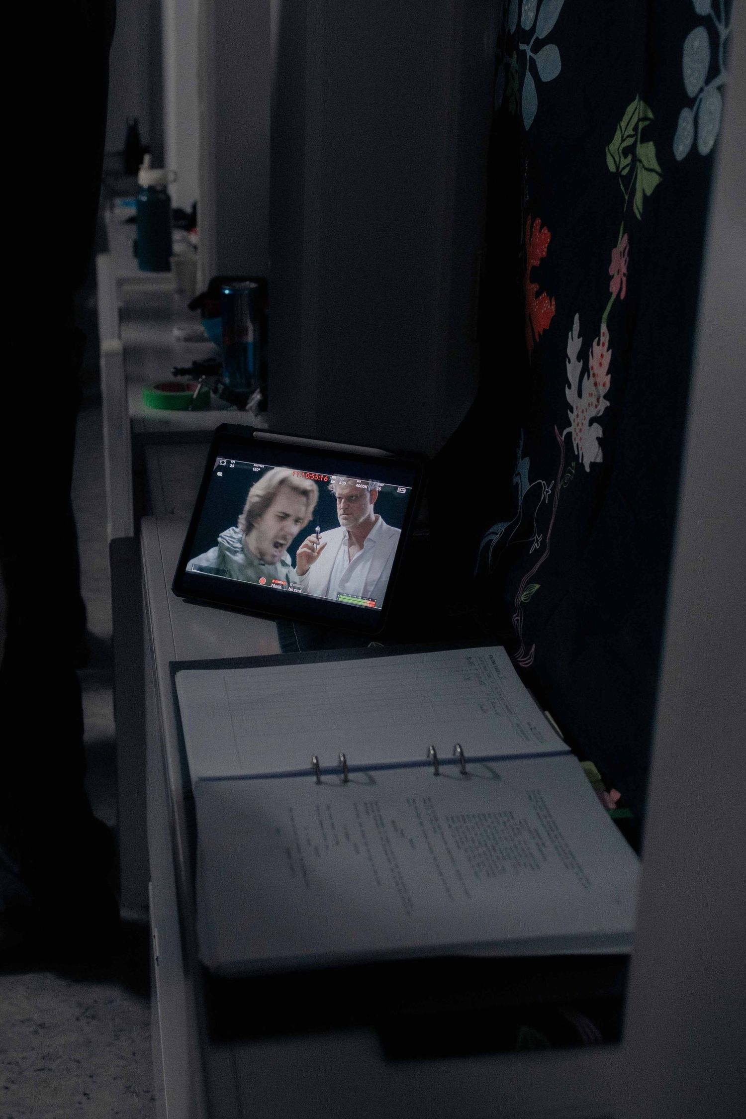 Script binder and tablet monitor displaying actors on a dark film production set backstage.