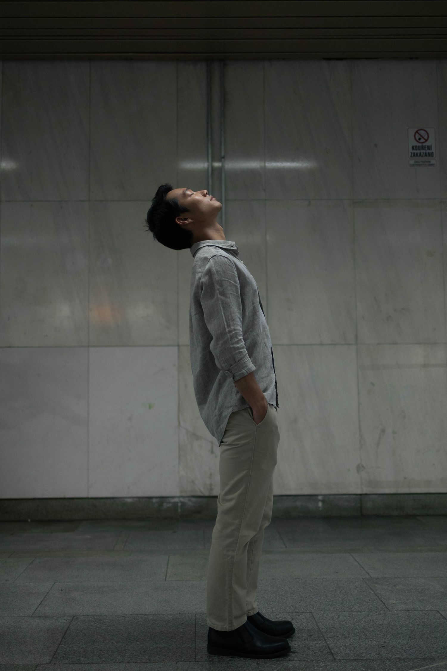 Young man in grey shirt tilts head back while standing in dimly lit subway station.