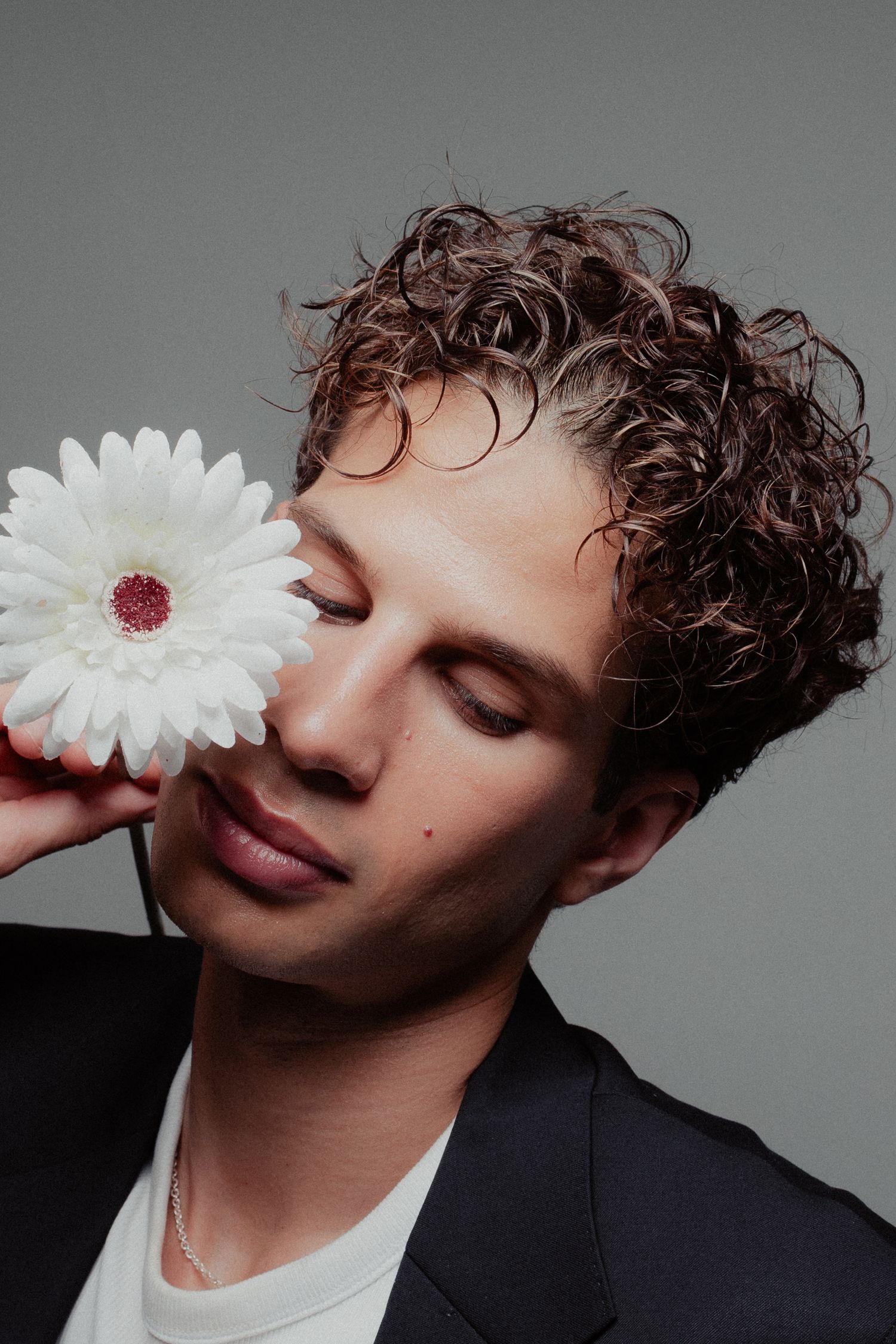 Young man with curly hair holding a white gerbera daisy over his eye in a studio portrait.