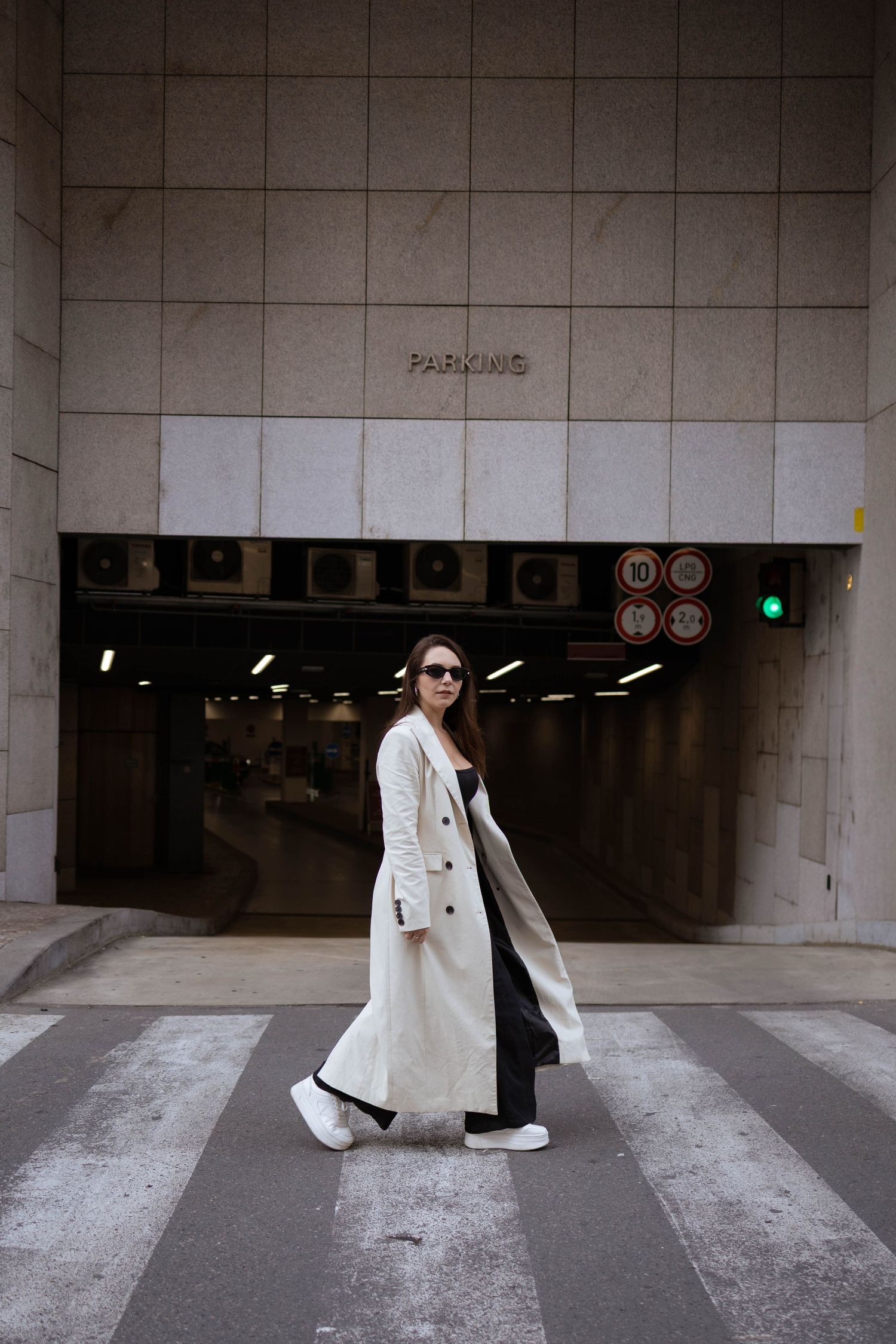 A stylish woman in a long white coat and sunglasses crosses a zebra crossing near a parking entrance.