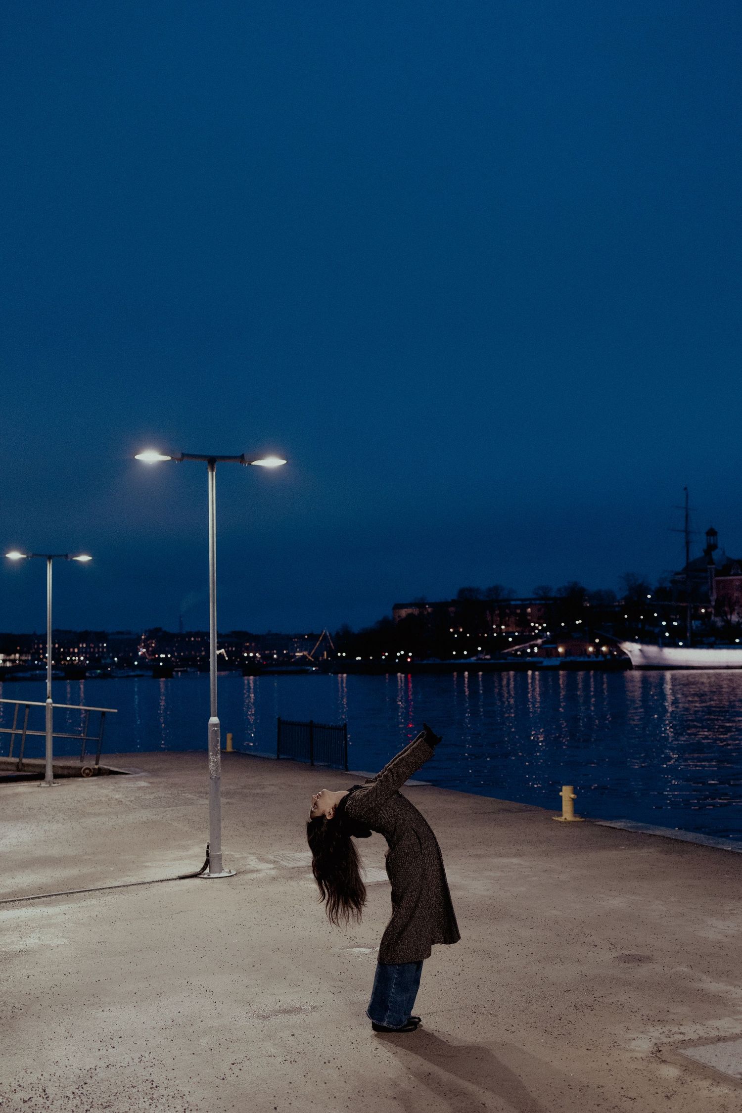 Woman bending backward dramatically near harbor street lamps at blue dusk.