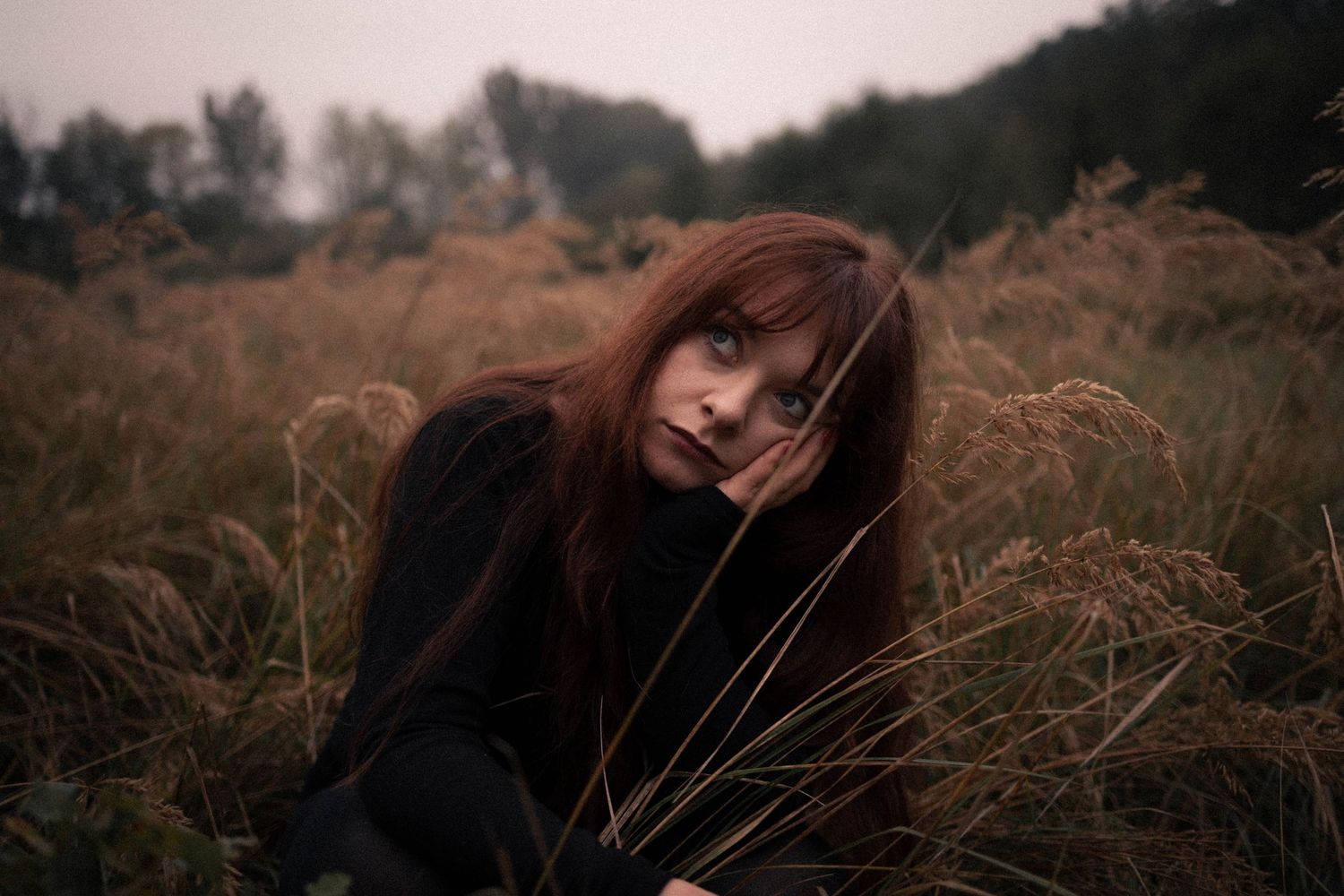 A young woman with red hair sits among tall dry grass in a moody autumn field.