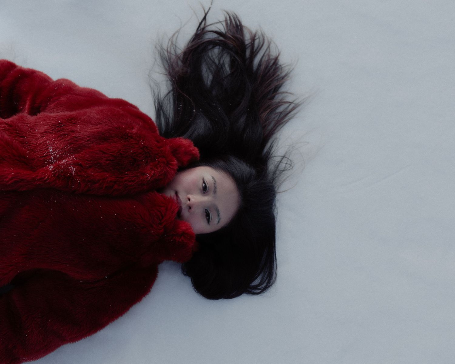 Woman in red coat lying in snow with dark hair spread around her.