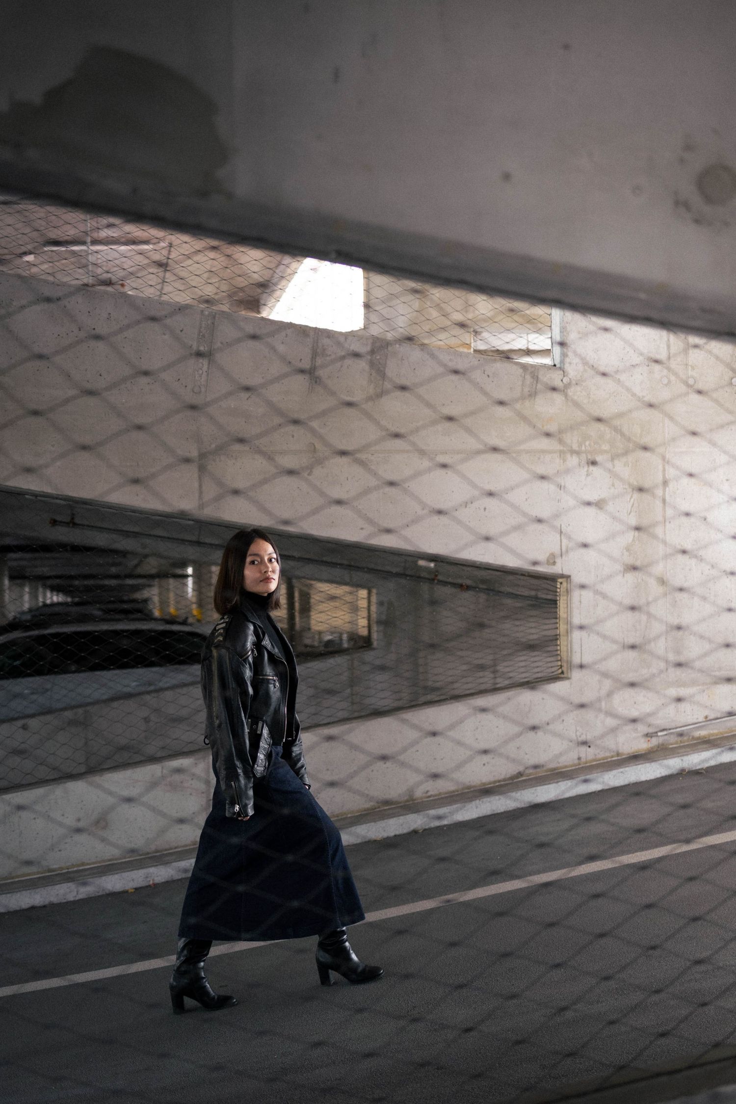 A woman in a black leather jacket and long skirt walks through an urban parking structure setting.