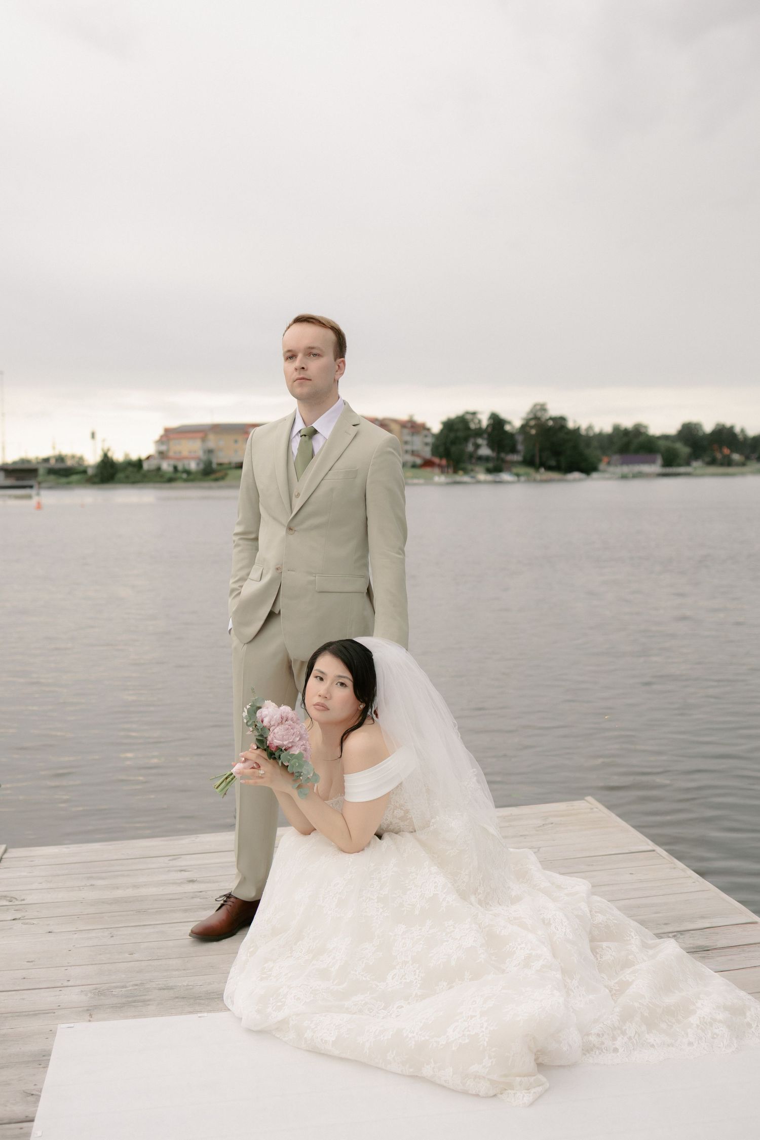 Wedding couple on dock by lake, groom standing in tan suit, bride seated.