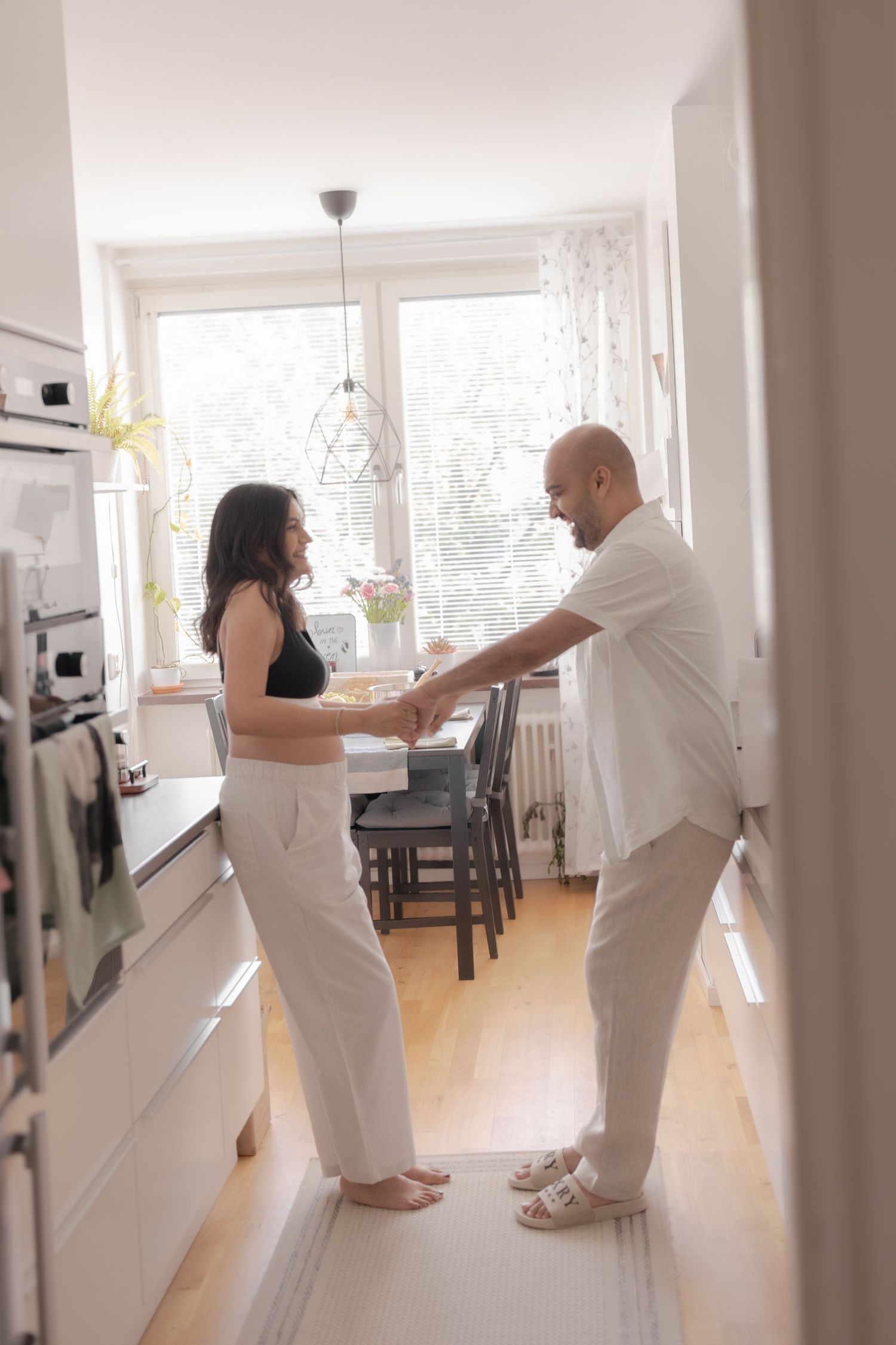 Couple holding hands and dancing together in a bright white modern kitchen with large windows.