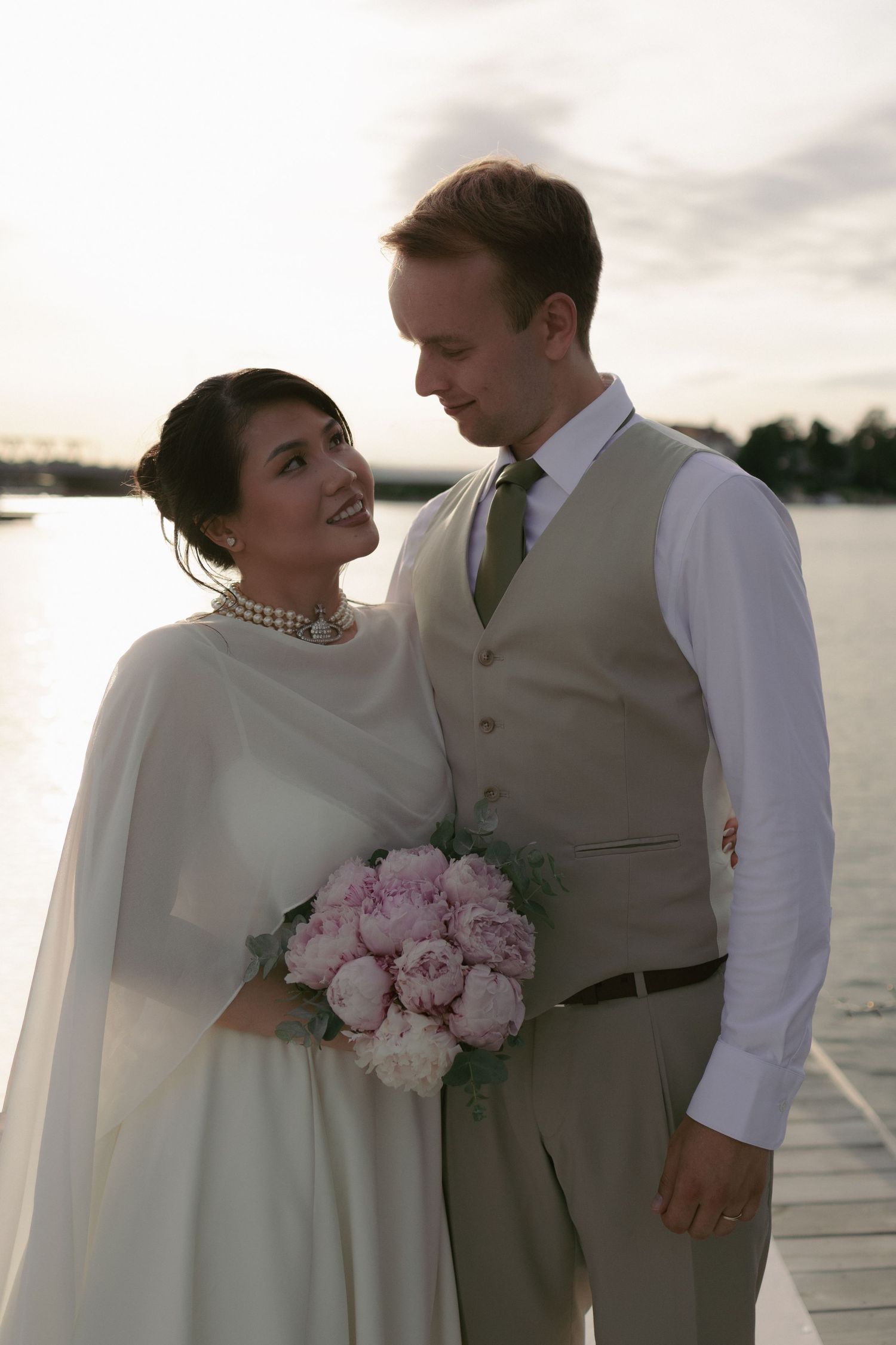 Bride in white cape holding pink peony bouquet gazes at groom in beige vest on waterfront dock at dusk.
