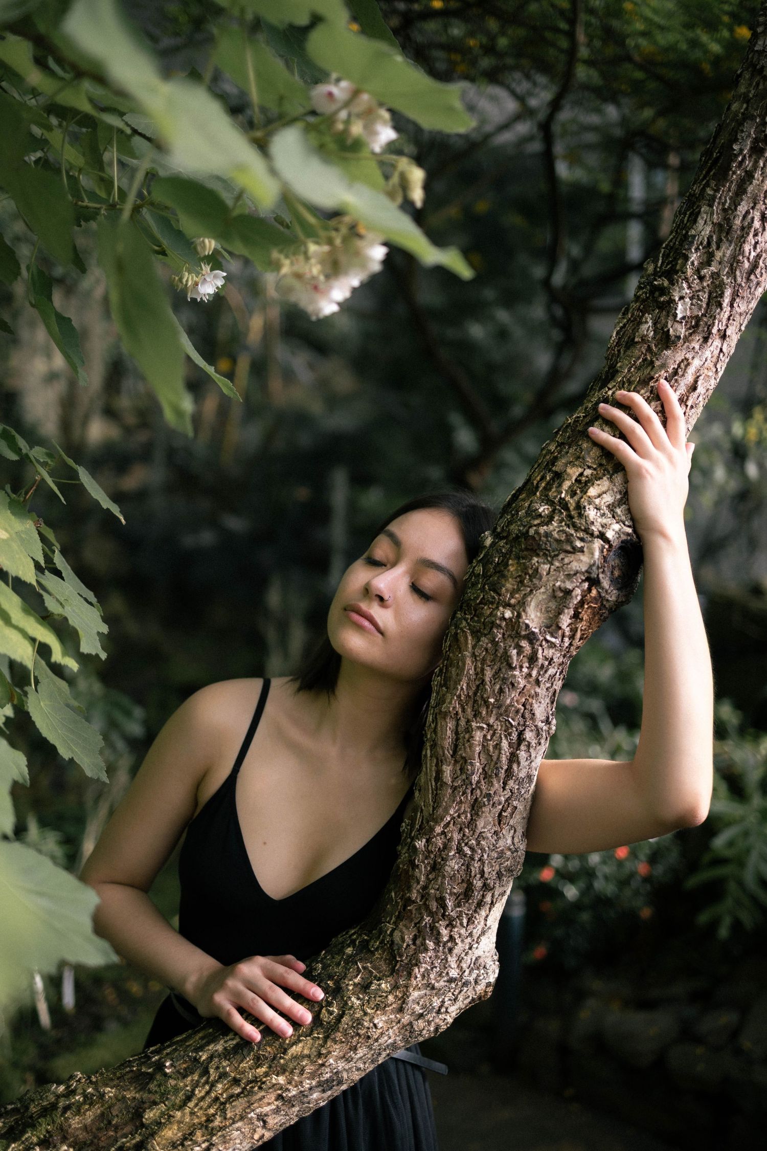 A serene young woman in a black dress leans against a tree trunk with eyes closed in a lush garden.