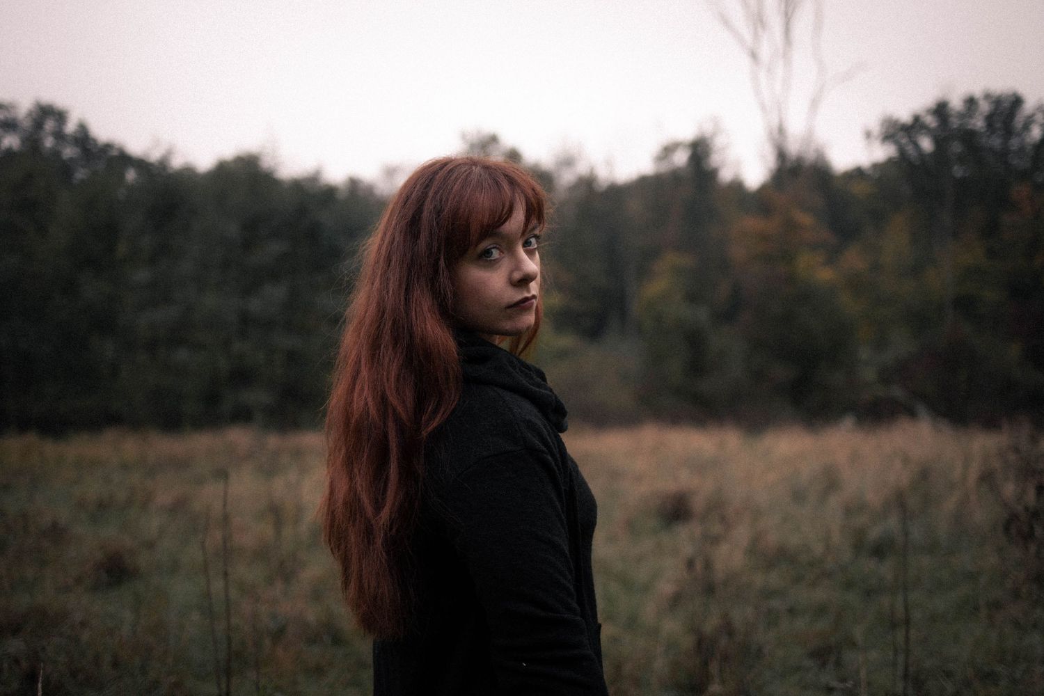 A young woman with long red hair stands in a field wearing a black top against a moody autumn backdrop.