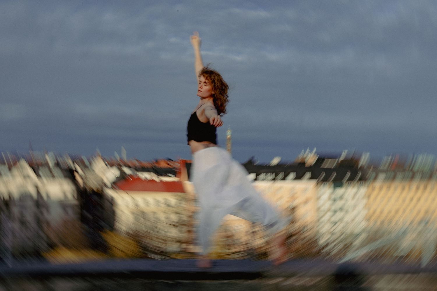A woman in a crop top and white pants jumps dynamically on a rooftop with a blurred cityscape behind.