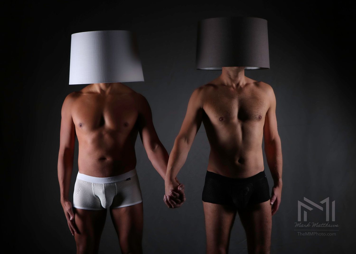 Two men in underwear hold hands with lampshades covering their heads in creative artistic photography.