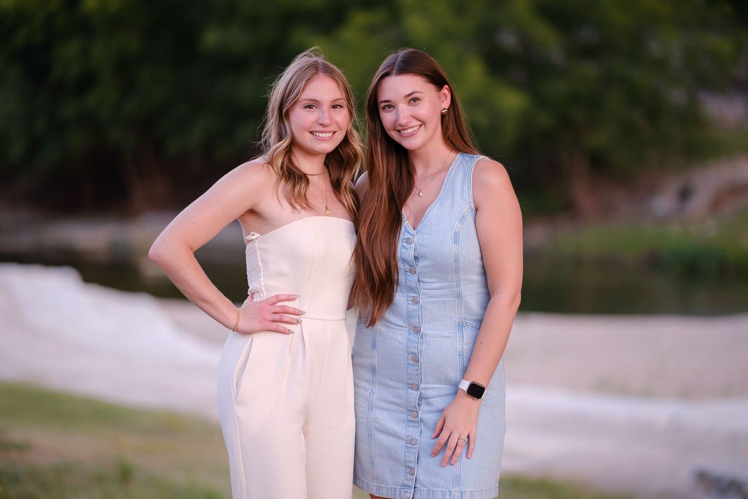 Two smiling young women in summer outfits posing together outdoors near water.