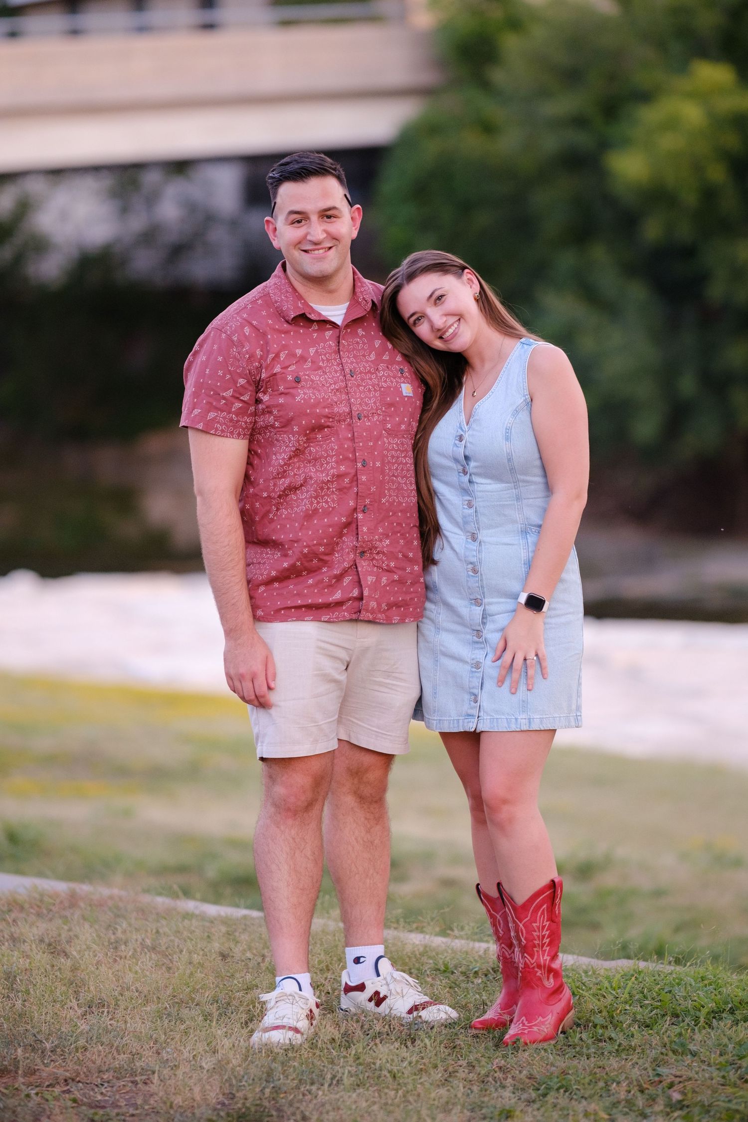 Smiling couple posing outdoors, man in red shirt and woman in denim dress with cowboy boots.