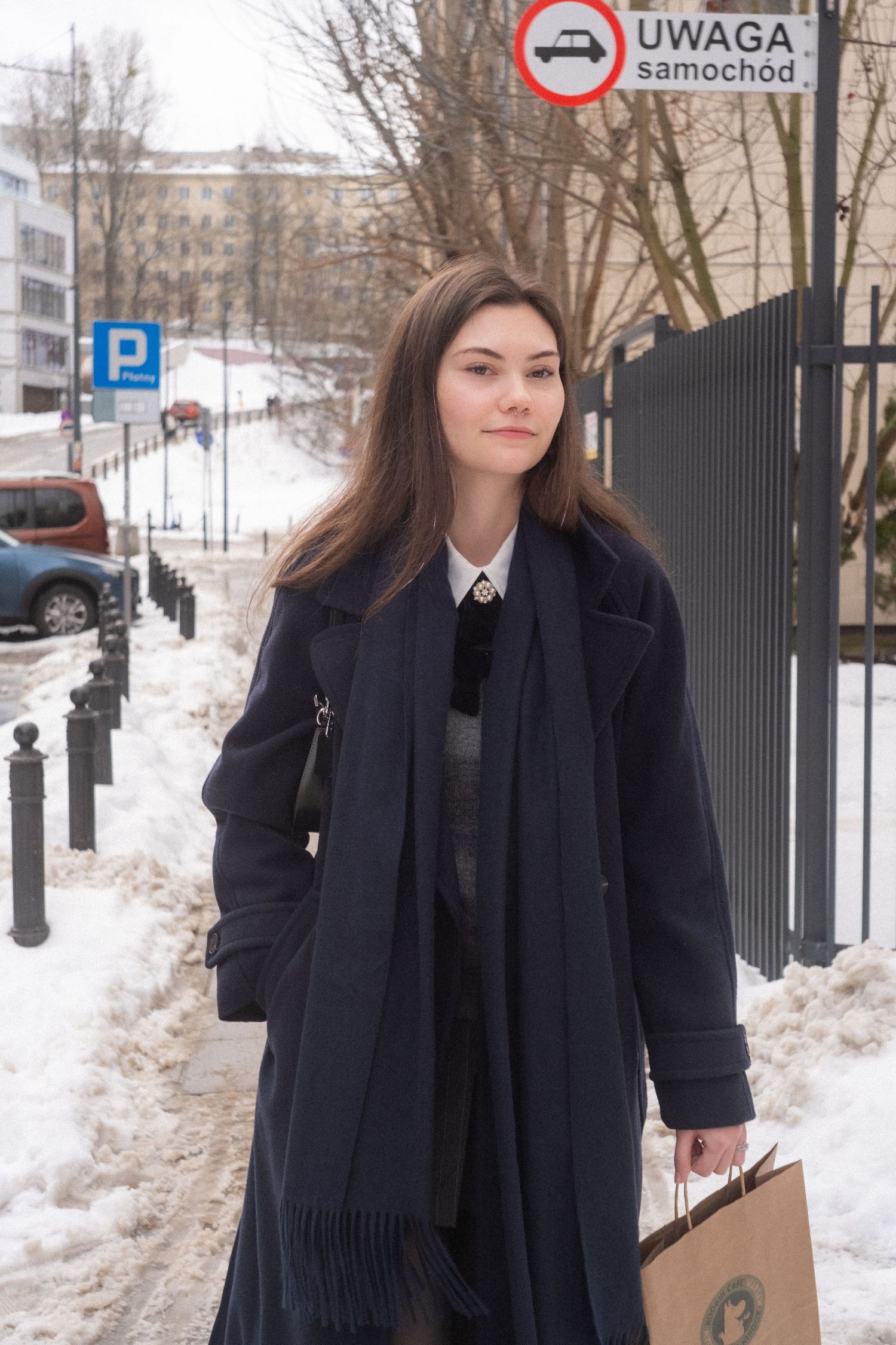 Young woman in navy coat carrying shopping bag on snowy city sidewalk in Poland.