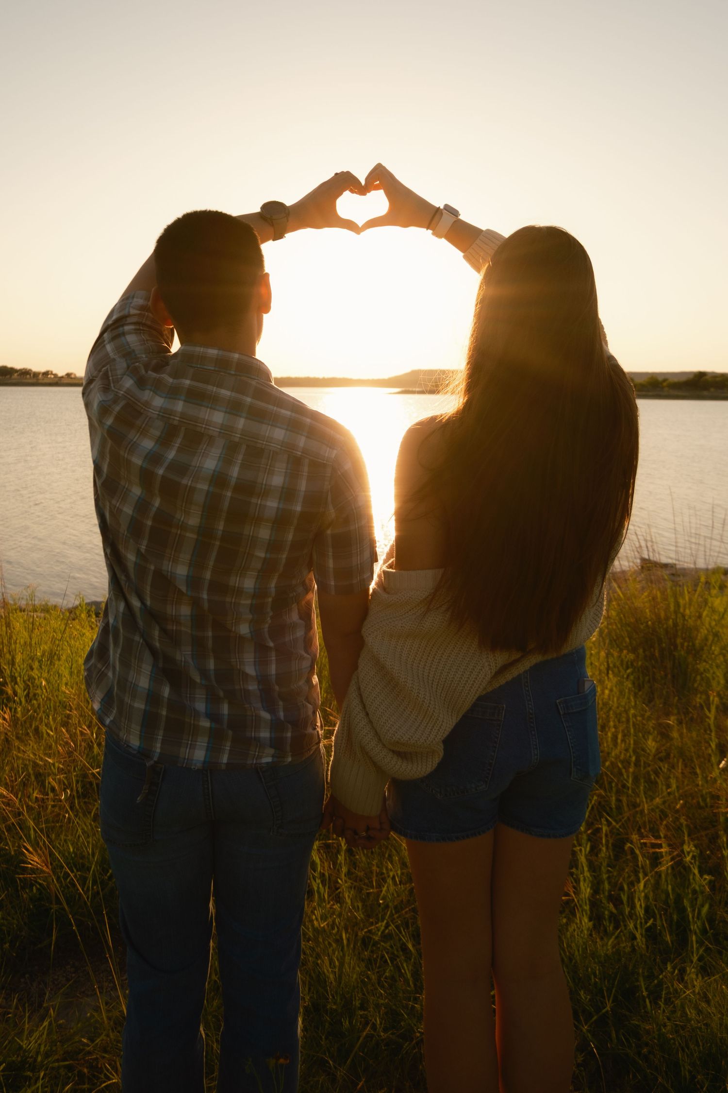 A couple makes a heart shape with their hands framing the sunset over a tranquil lake.