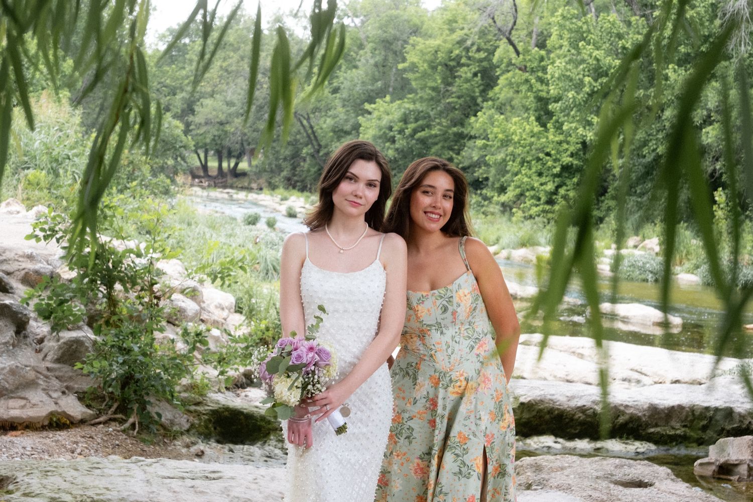 Two young women in summer dresses pose together in a lush outdoor garden setting with flowers.