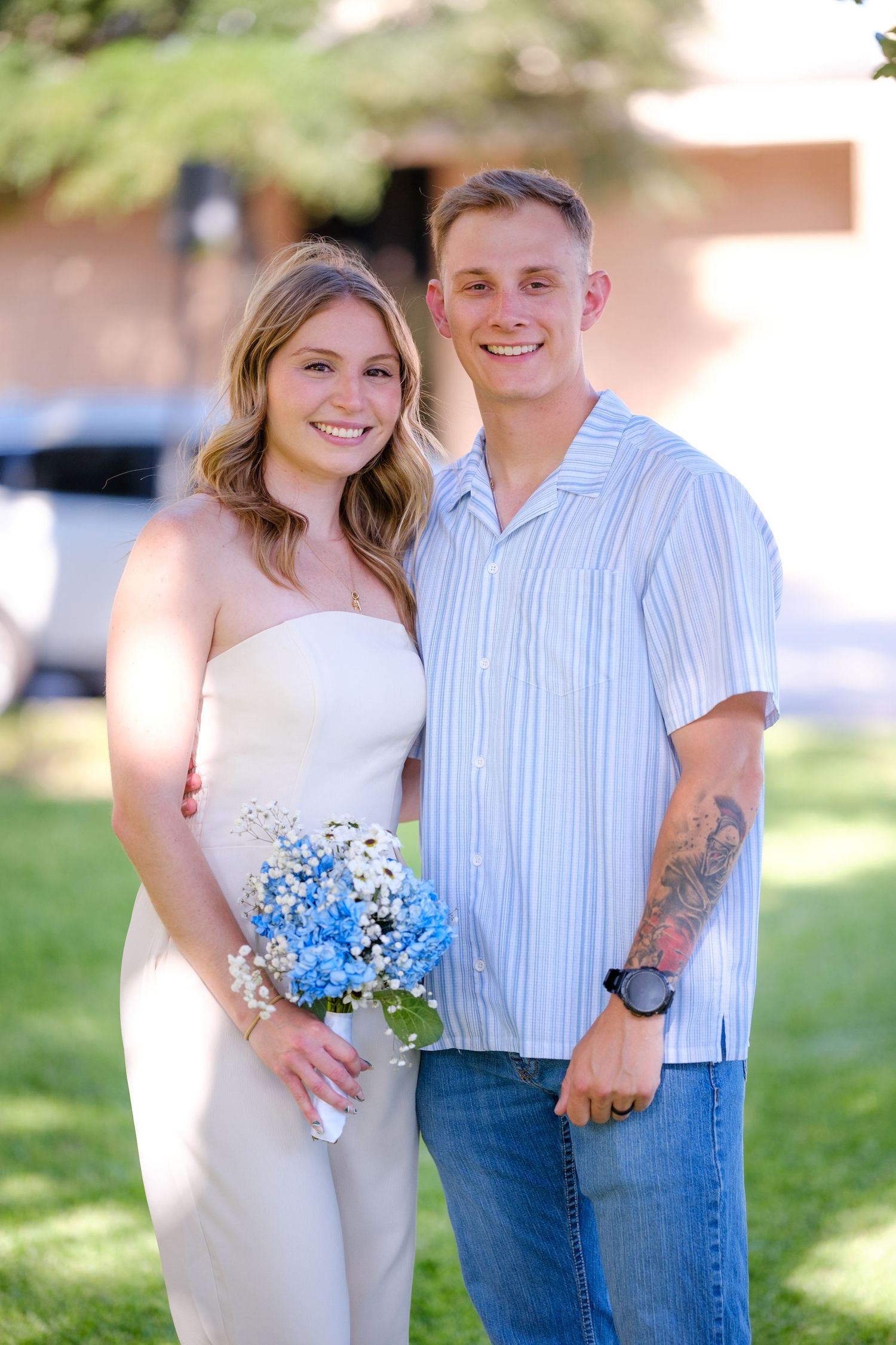 A smiling couple poses outdoors, the woman holding a bouquet of blue and white flowers.
