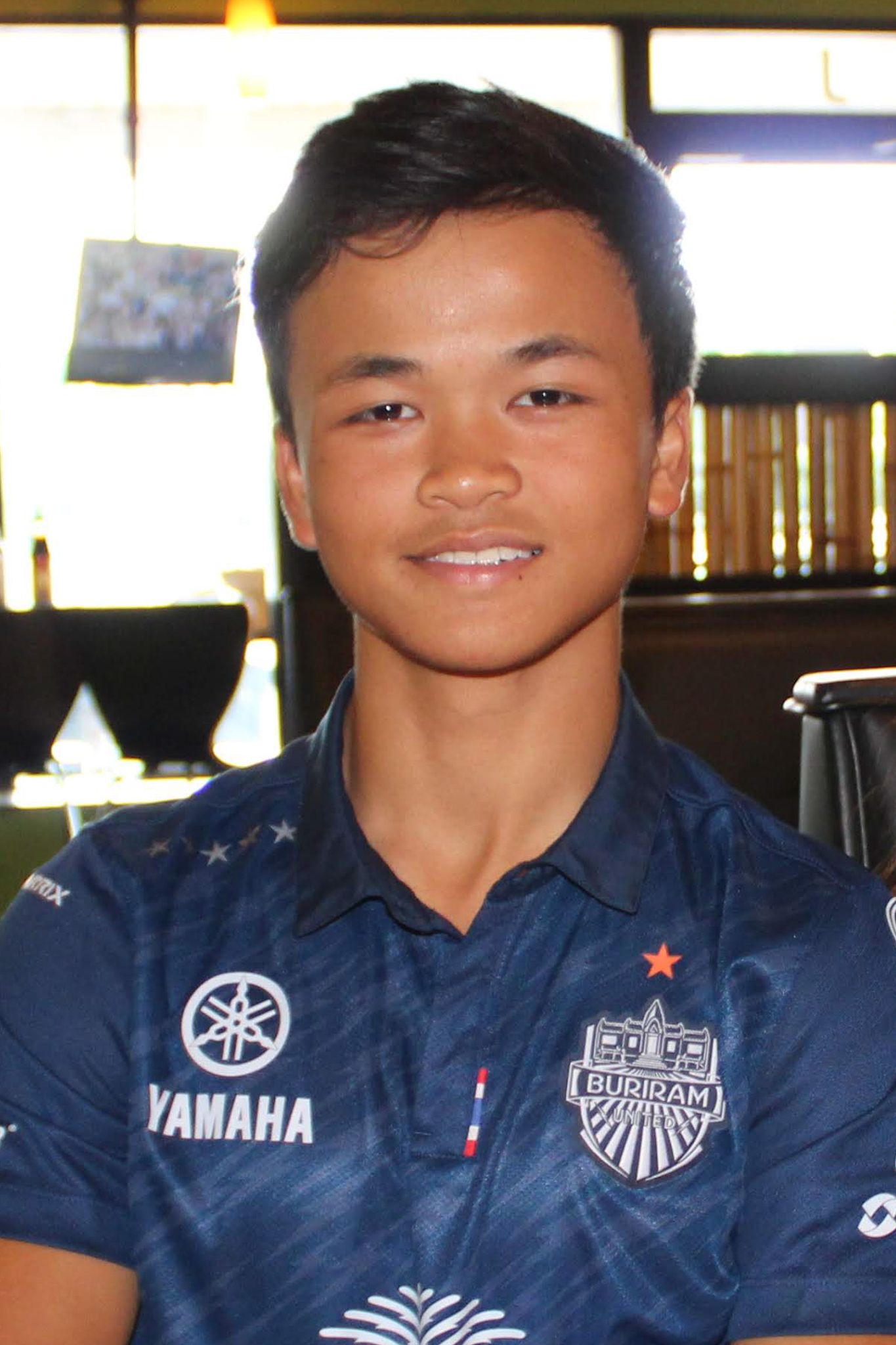 A young man smiles wearing a Yamaha Buridam FC soccer jersey inside a restaurant.