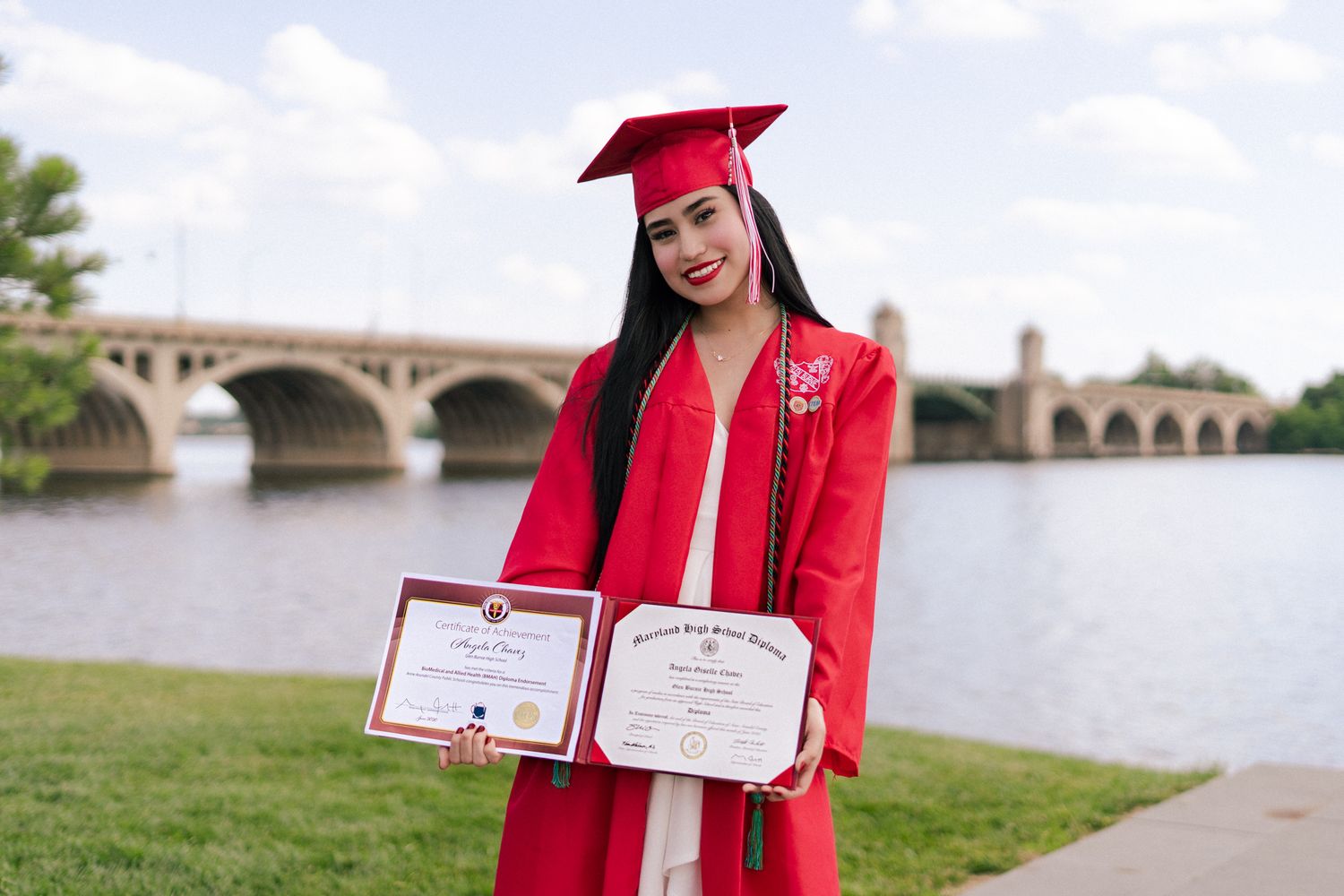 Smiling female graduate in red cap and gown holding two diplomas near a river bridge.