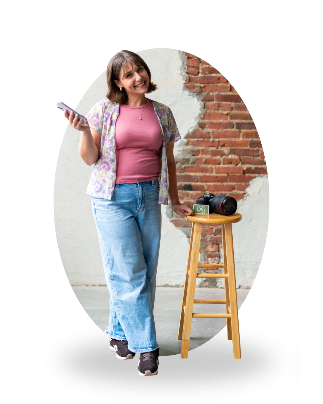 Layla wearing a pink top and jeans, leaning against brick wall holding phone, with camera on wooden stool beside her.