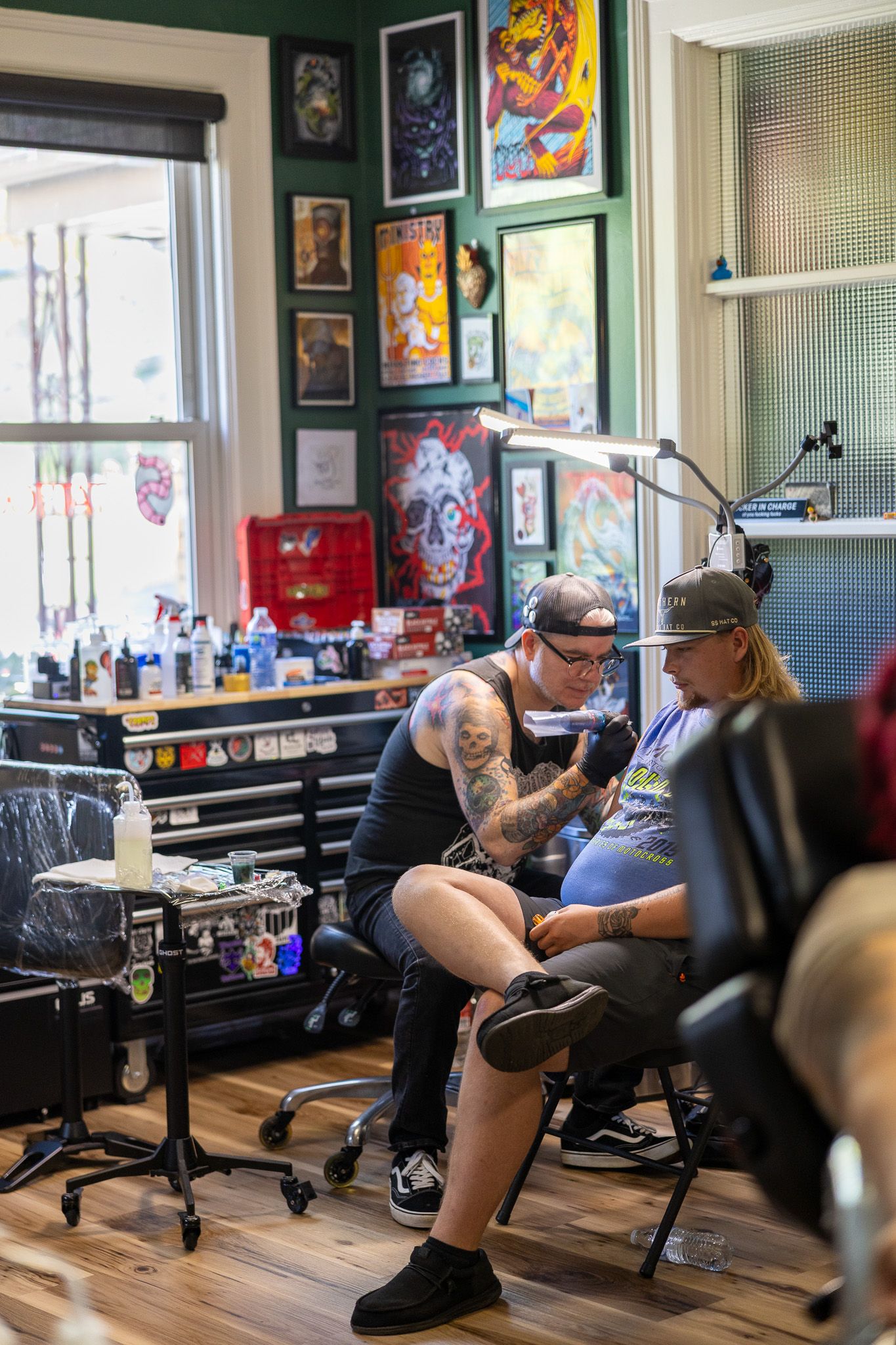 Tattoo artist with sleeve tattoos working on client in vibrant studio with colorful art-covered green walls.