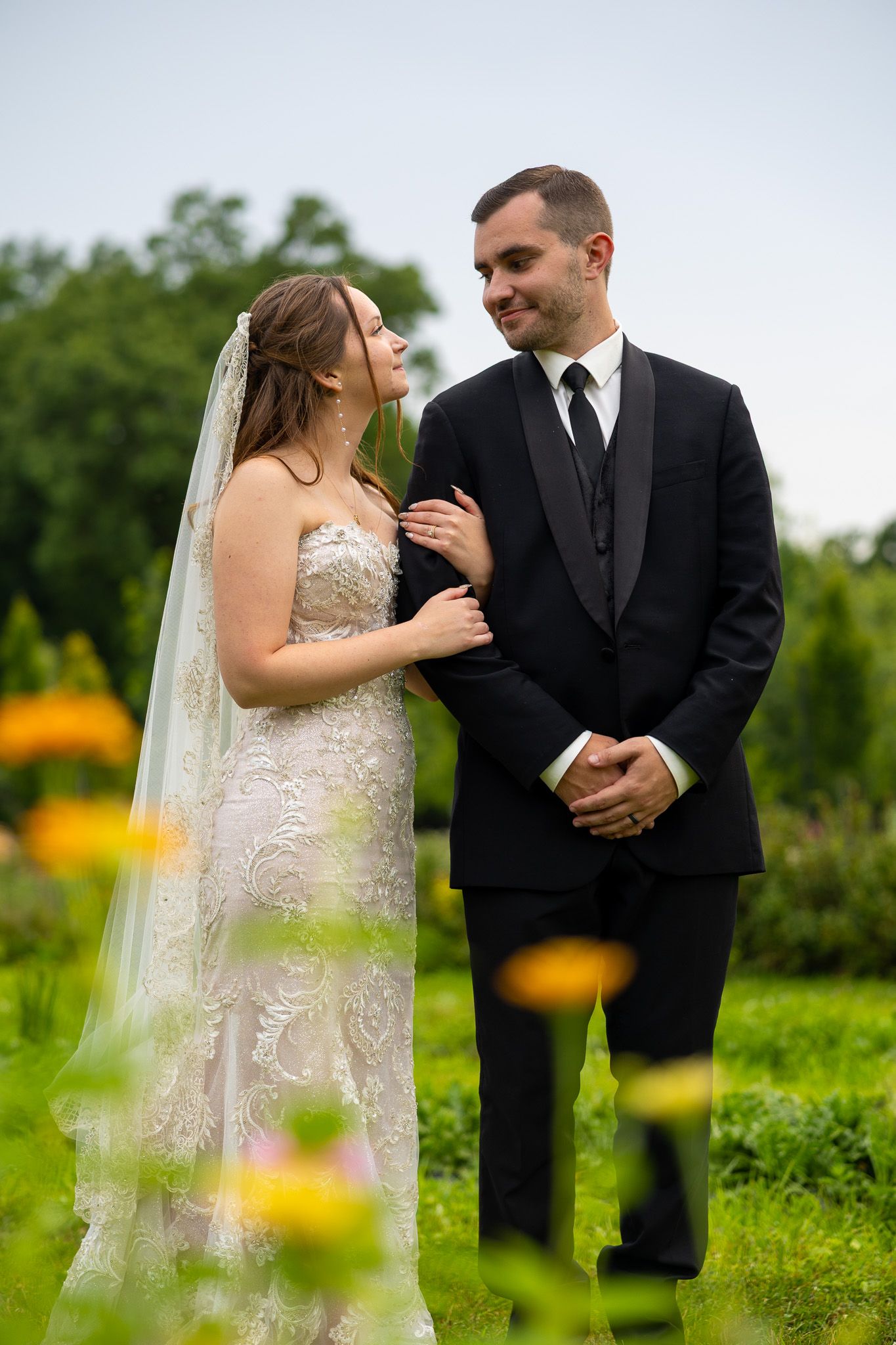 Bride and groom smiling at each other in garden with yellow flowers, groom in black tuxedo.