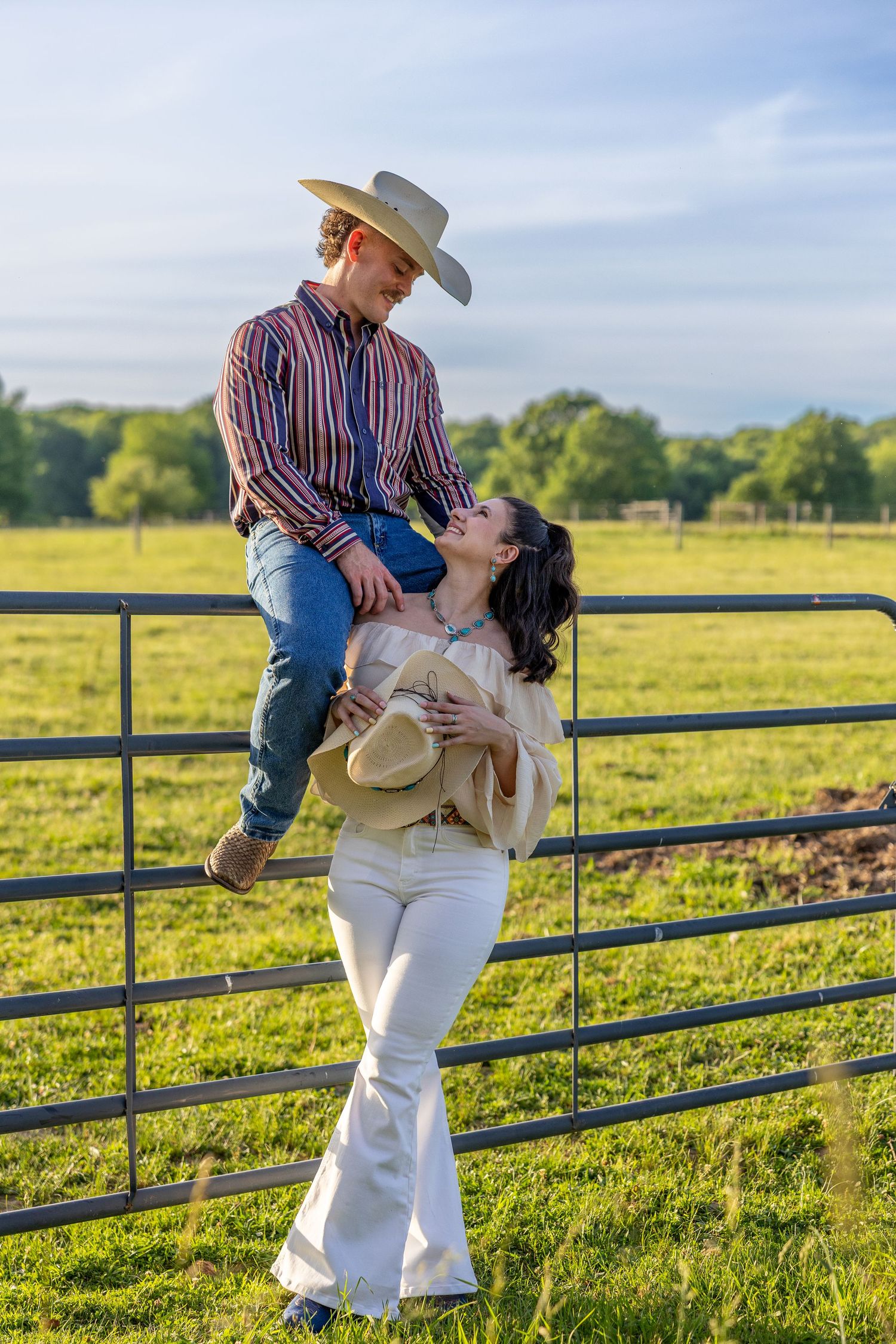 Man sitting on a farm gate leaning toward woman in white flare pants in a green field.