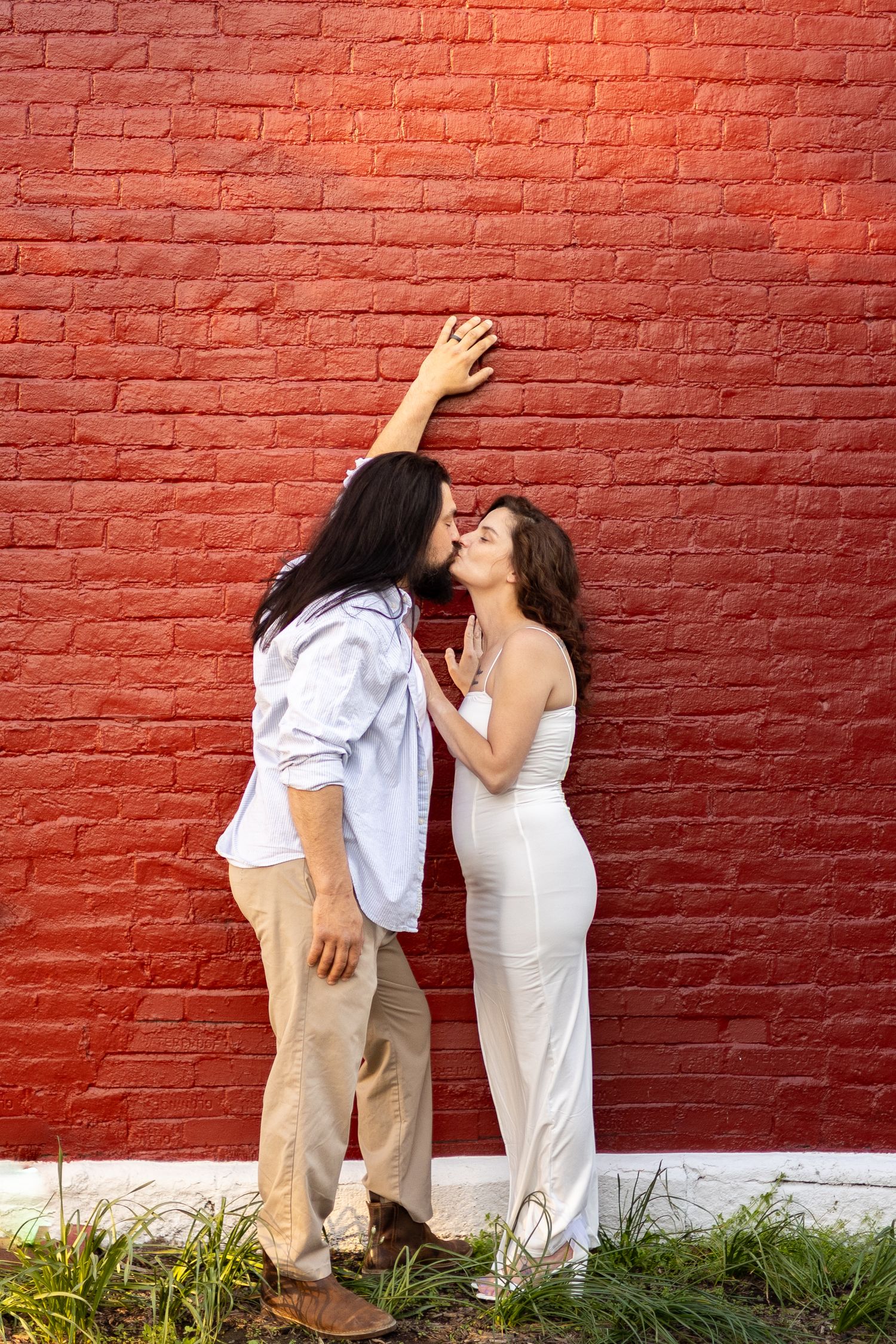 Couple sharing a kiss against a vibrant red brick wall outdoors.