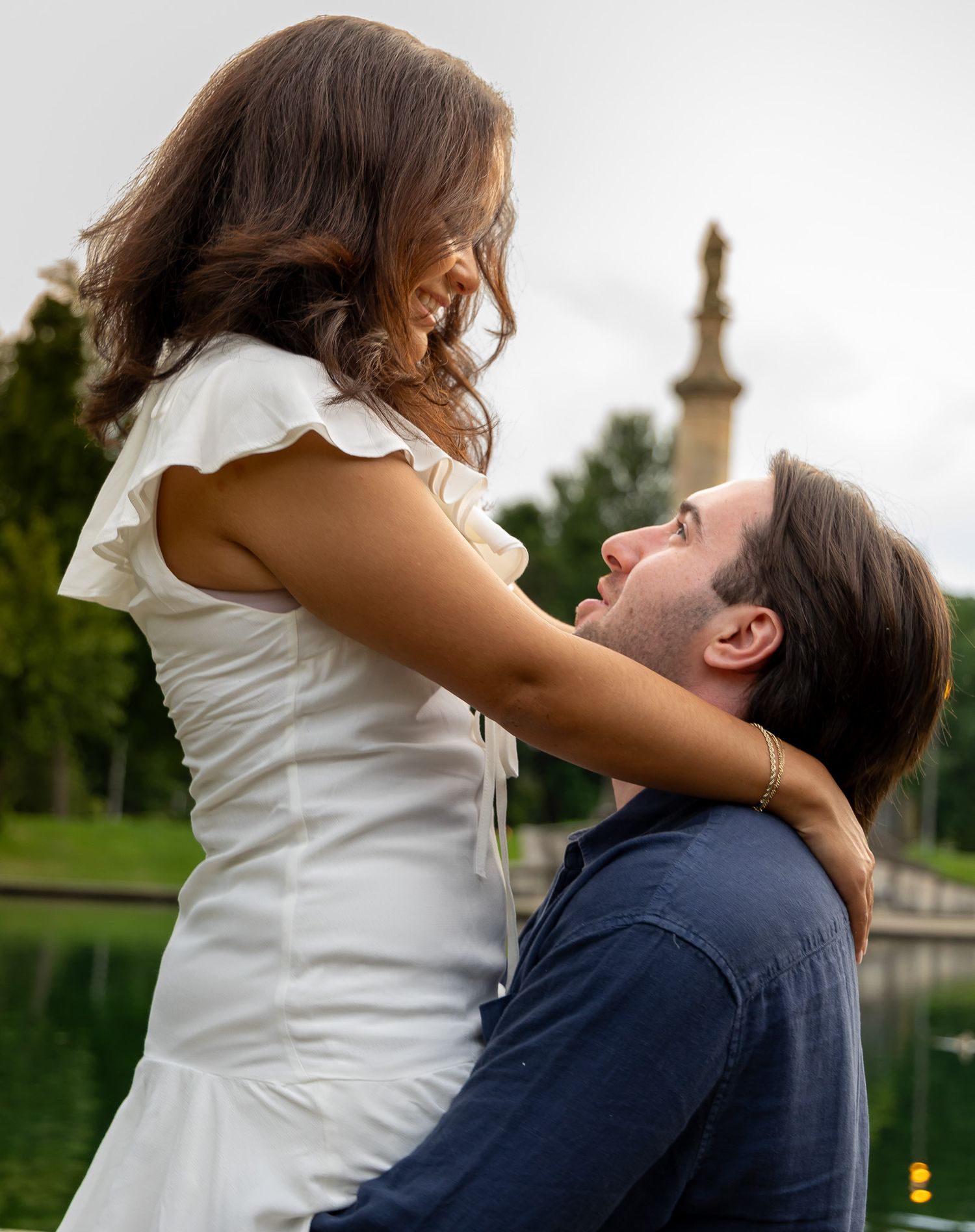 Couple sharing a romantic moment by a pond, woman in white dress smiling down at man in blue shirt.