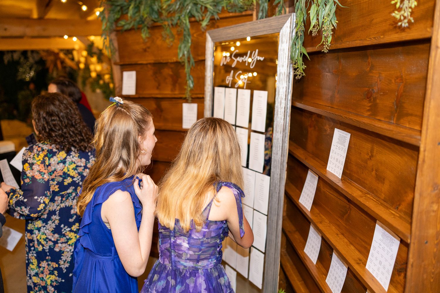 Wedding guests in blue dresses view seating chart displayed on rustic wooden wall at reception.