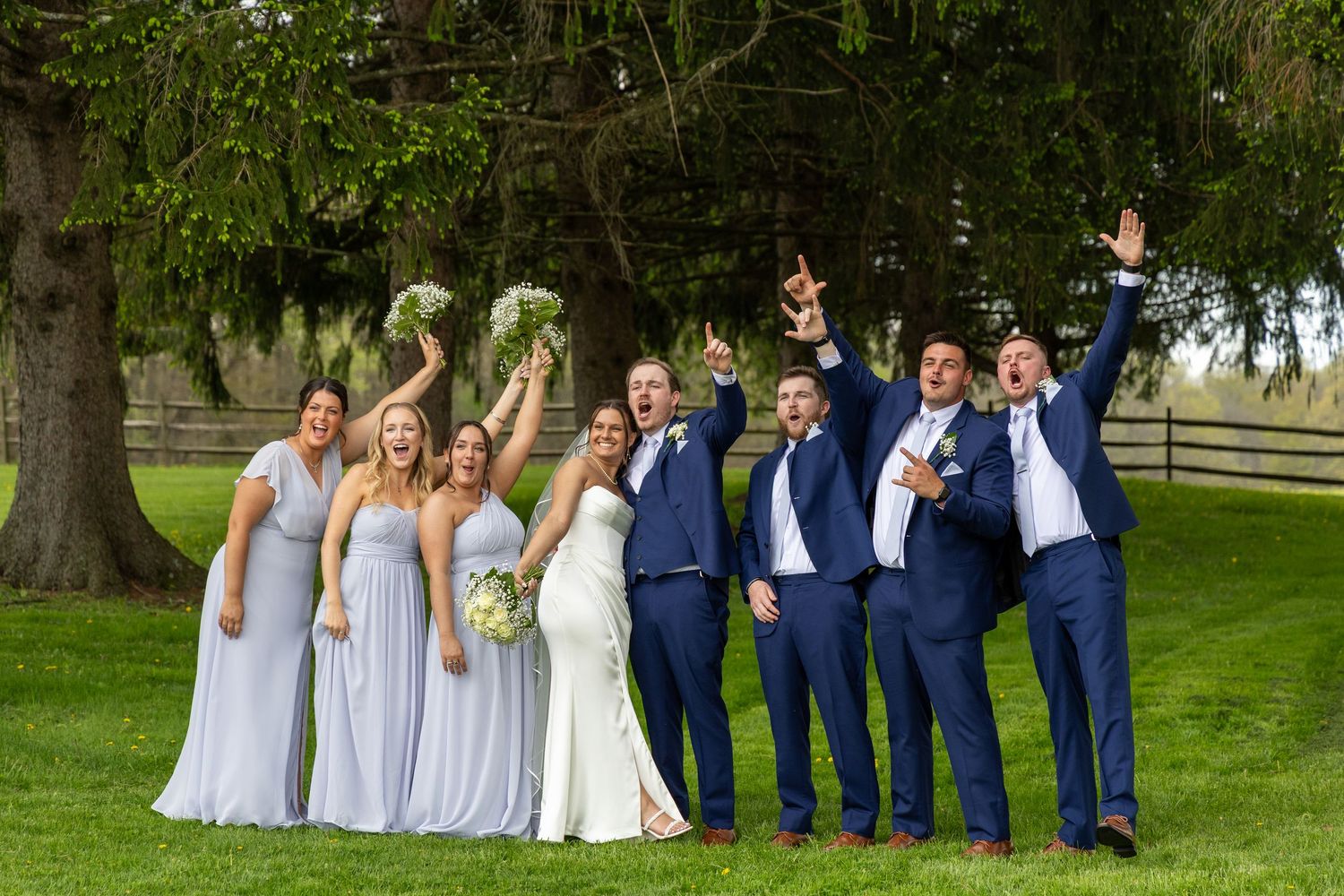 Joyful wedding party in blue suits and lavender gowns celebrating outdoors under a large tree.