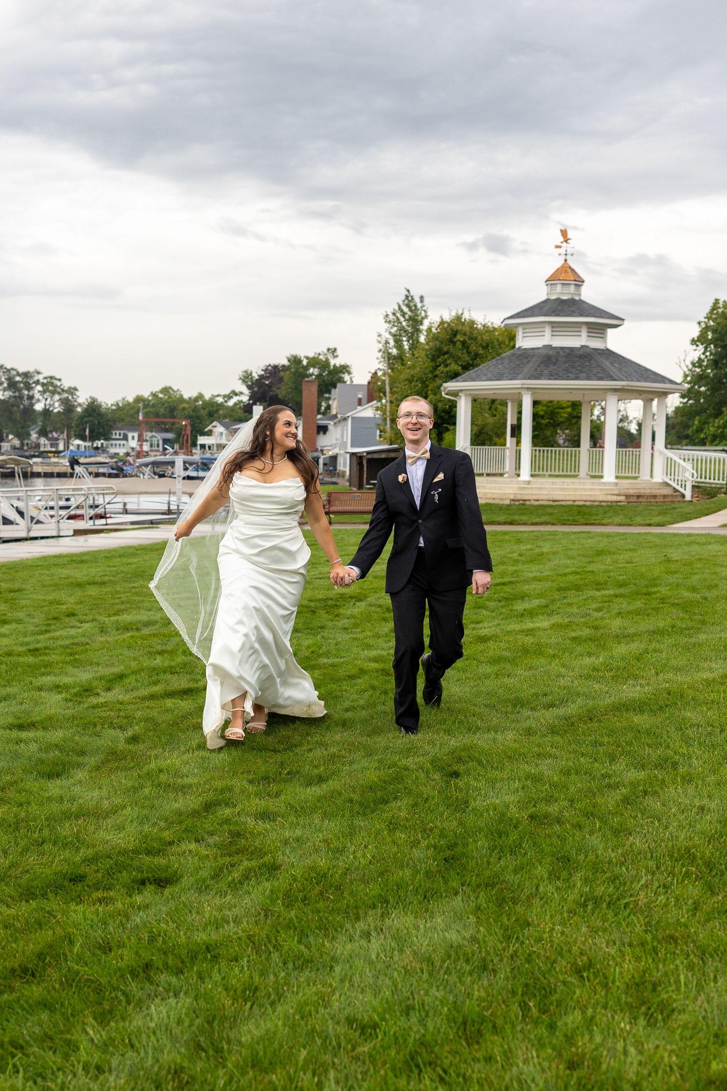 Bride and groom holding hands walking on green lawn near gazebo at waterfront wedding venue under cloudy sky.