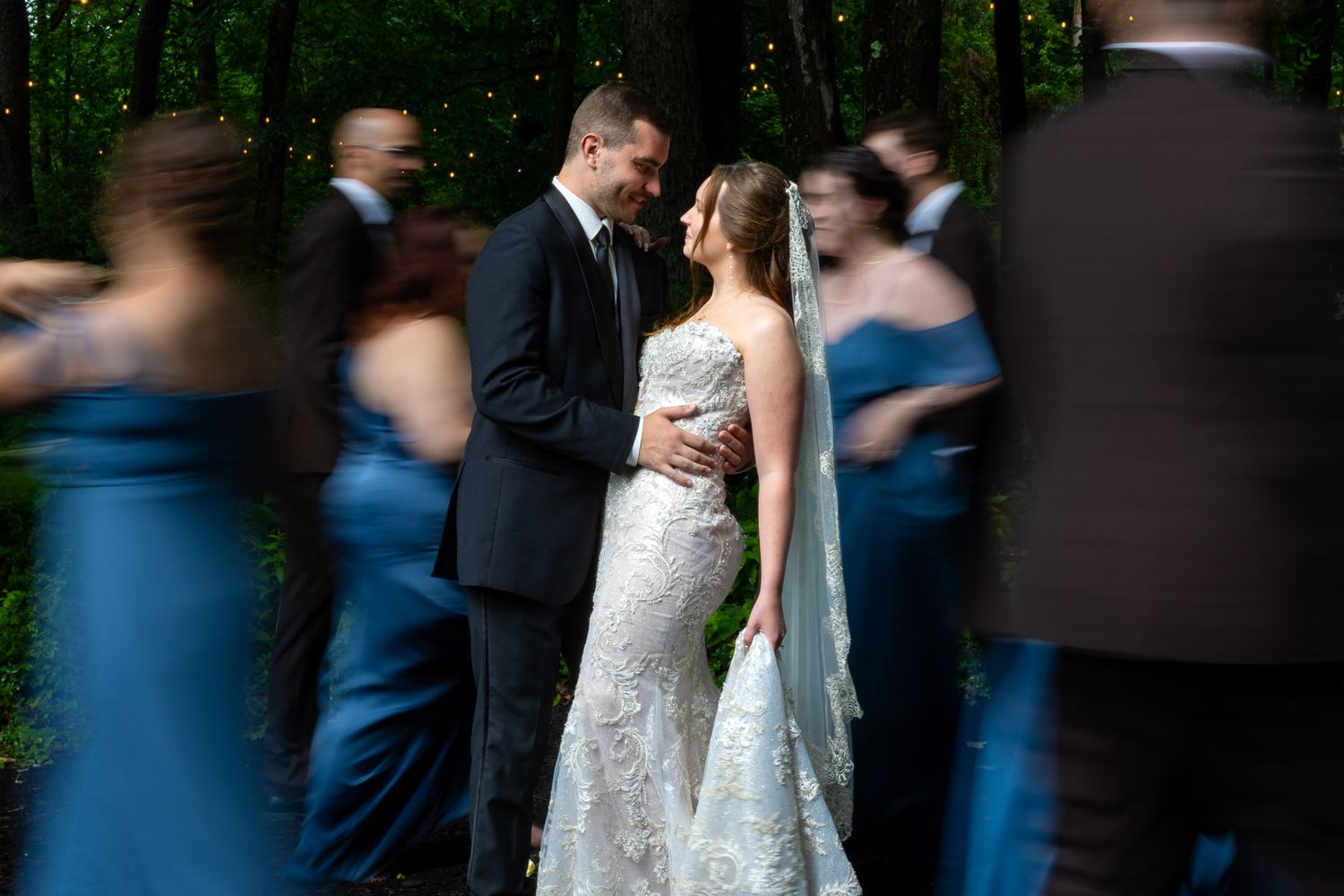 Bride and groom share an intimate moment surrounded by blurred wedding party in blue gowns in a forest setting.