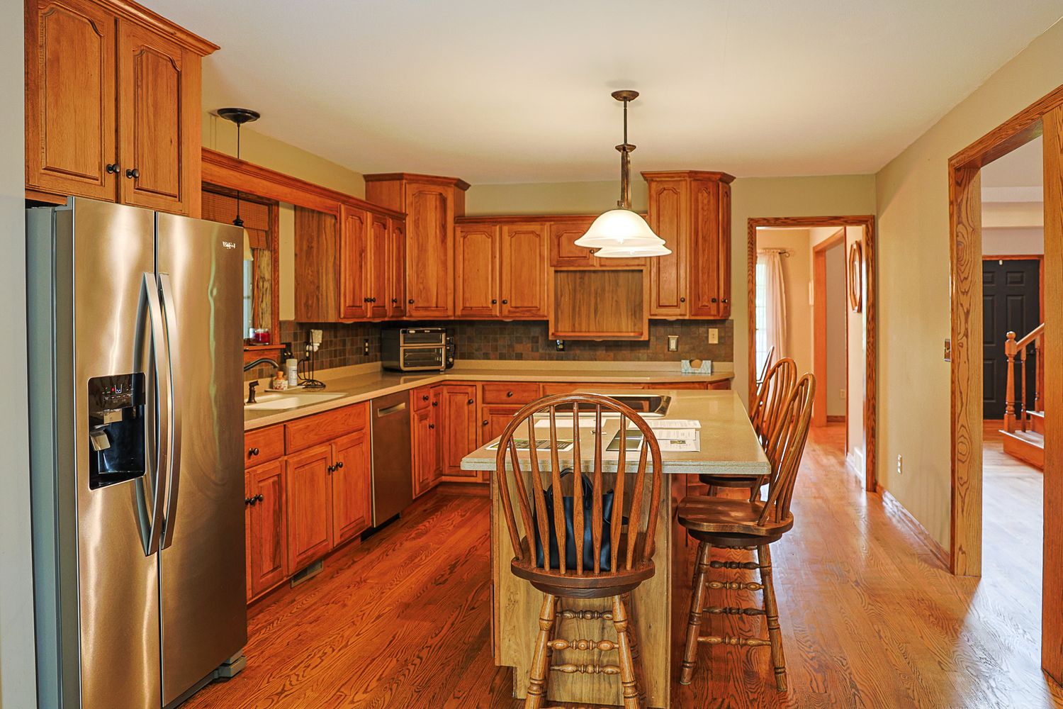 Spacious kitchen with oak cabinets, stainless steel fridge, hardwood floors, island with chairs, and pendant lighting.