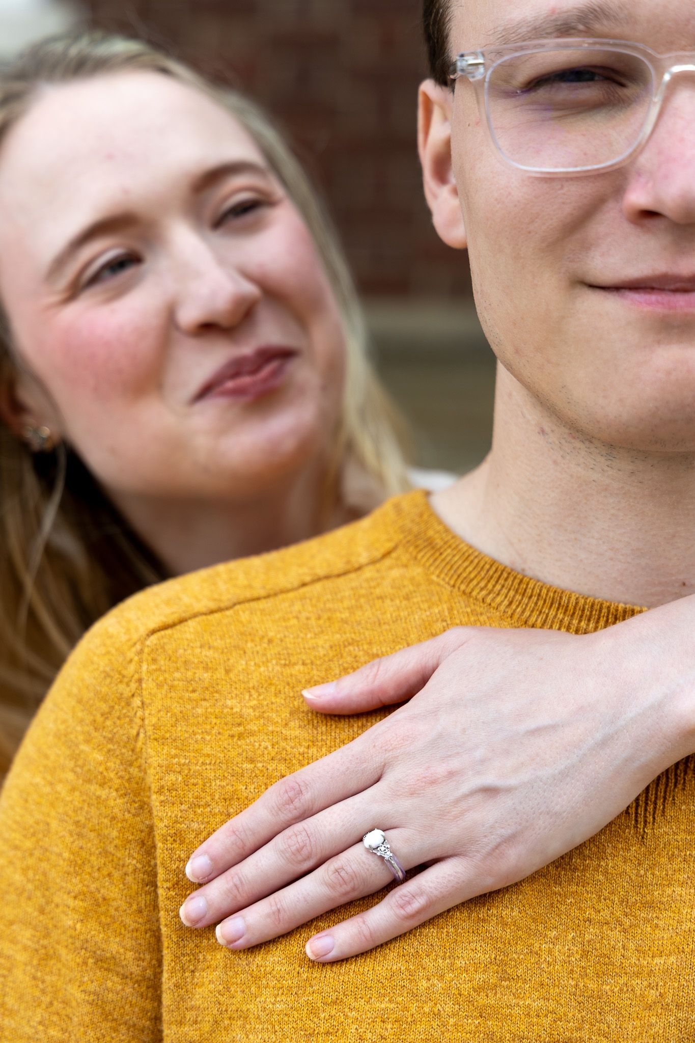 Couple in mustard sweaters, woman showing diamond engagement ring while making a kissing face behind her partner.