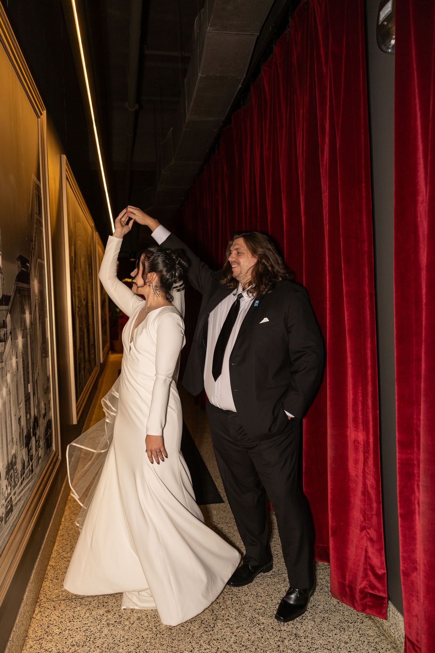 A bride in a white gown twirls as her groom in a black suit spins her near a red velvet curtain.