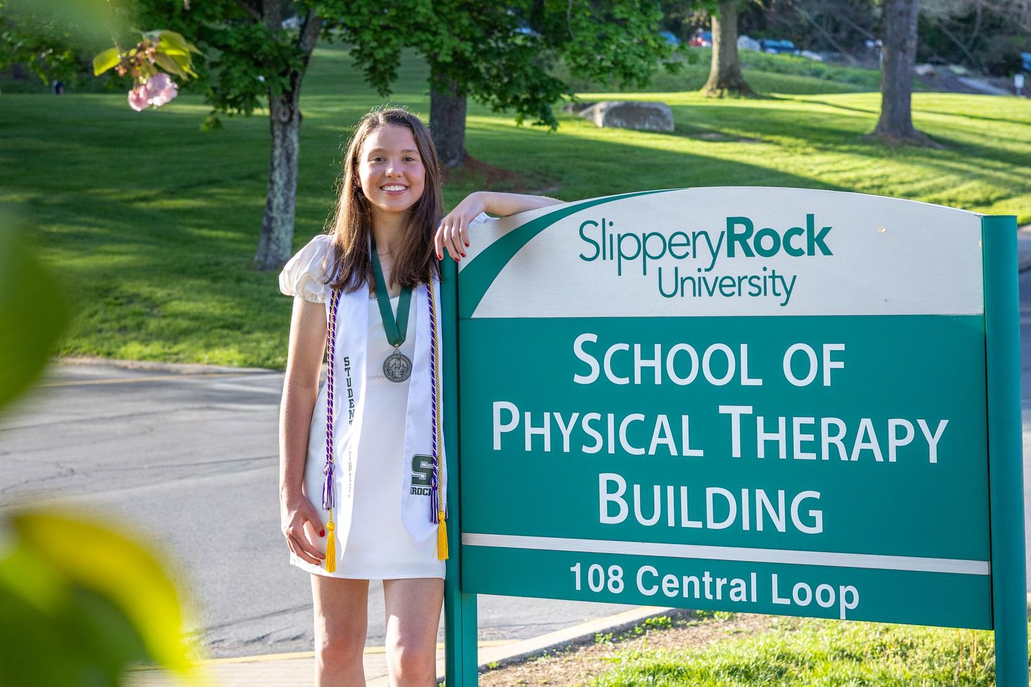 Graduate poses beside Slippery Rock University School of Physical Therapy Building sign with medal.