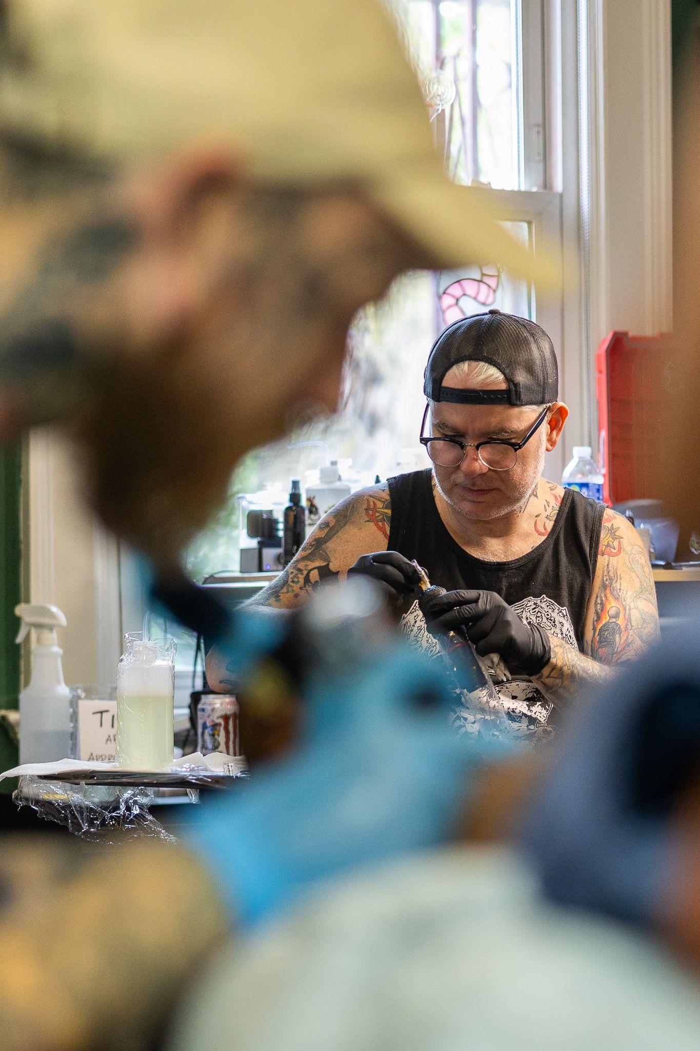 Heavily tattooed artist with backward cap and glasses tattooing a client in a bright studio.