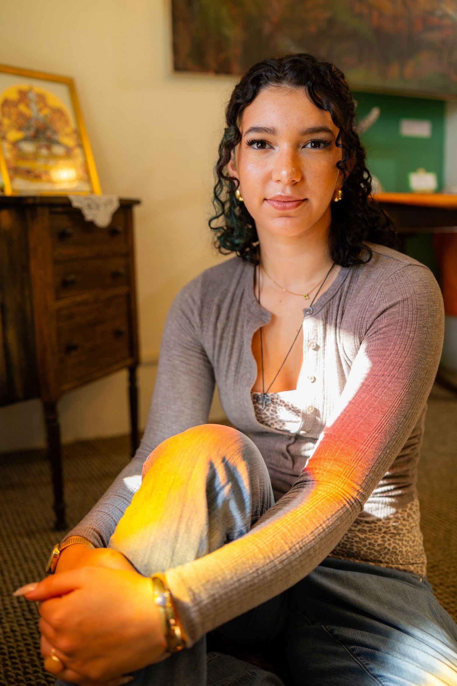 Young woman with curly hair sits cross-legged indoors, wearing gray cardigan, bathed in warm sunlight.
