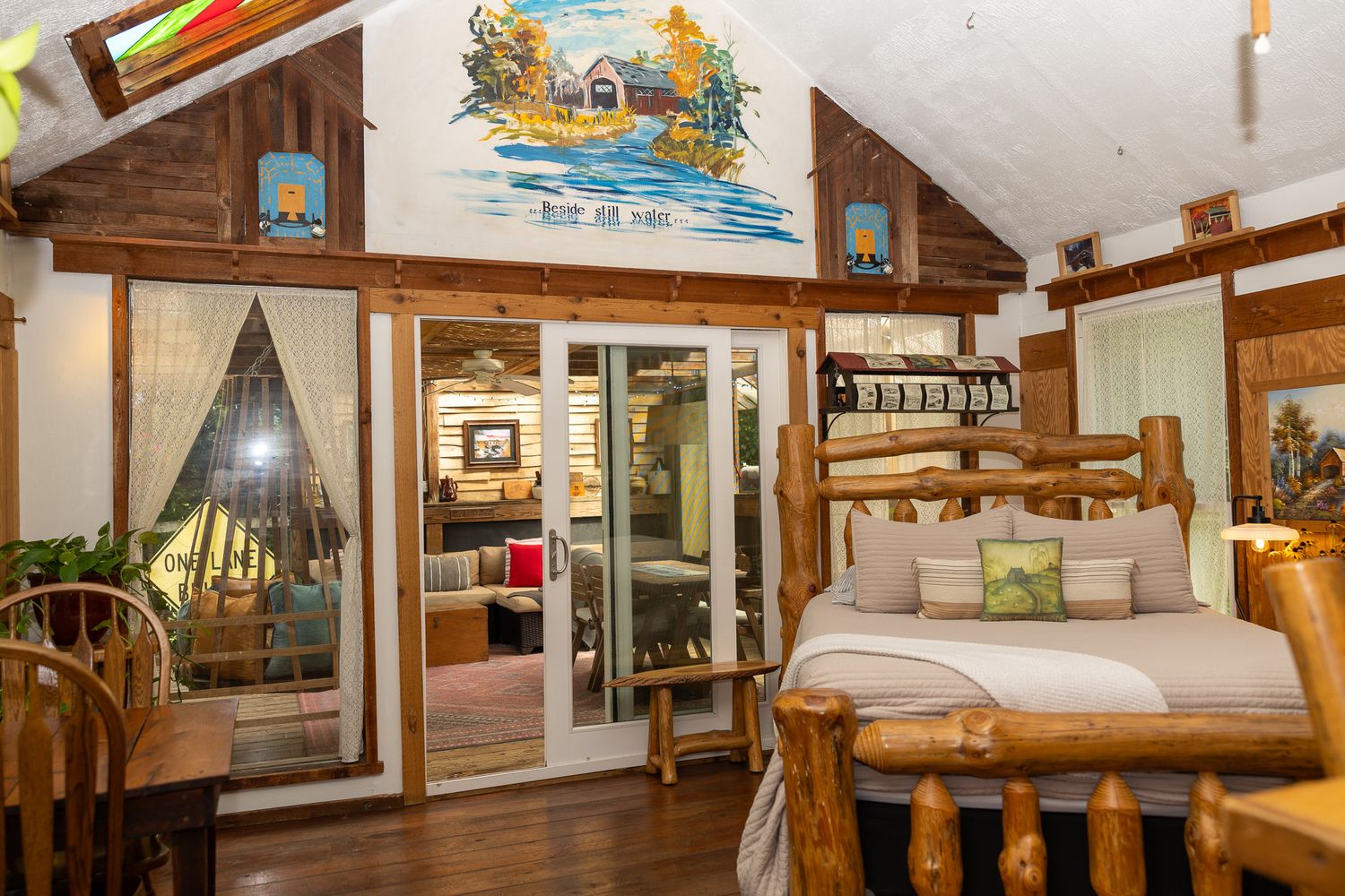 Rustic log cabin bedroom with bunk beds, vaulted ceiling, colorful mural, and sliding glass doors to living area.