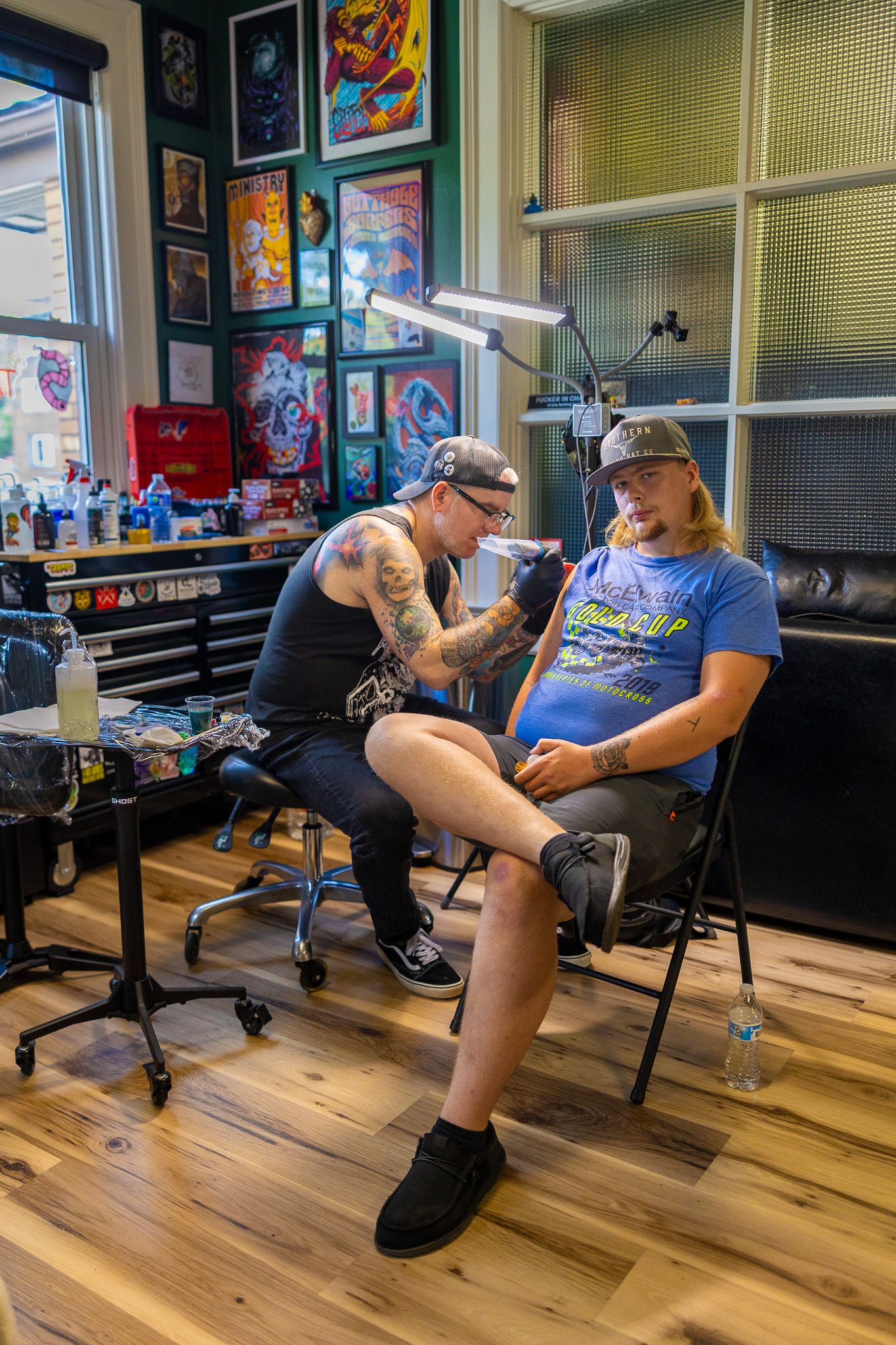 Tattoo artist applying ink to seated male client in bright studio filled with framed artwork.