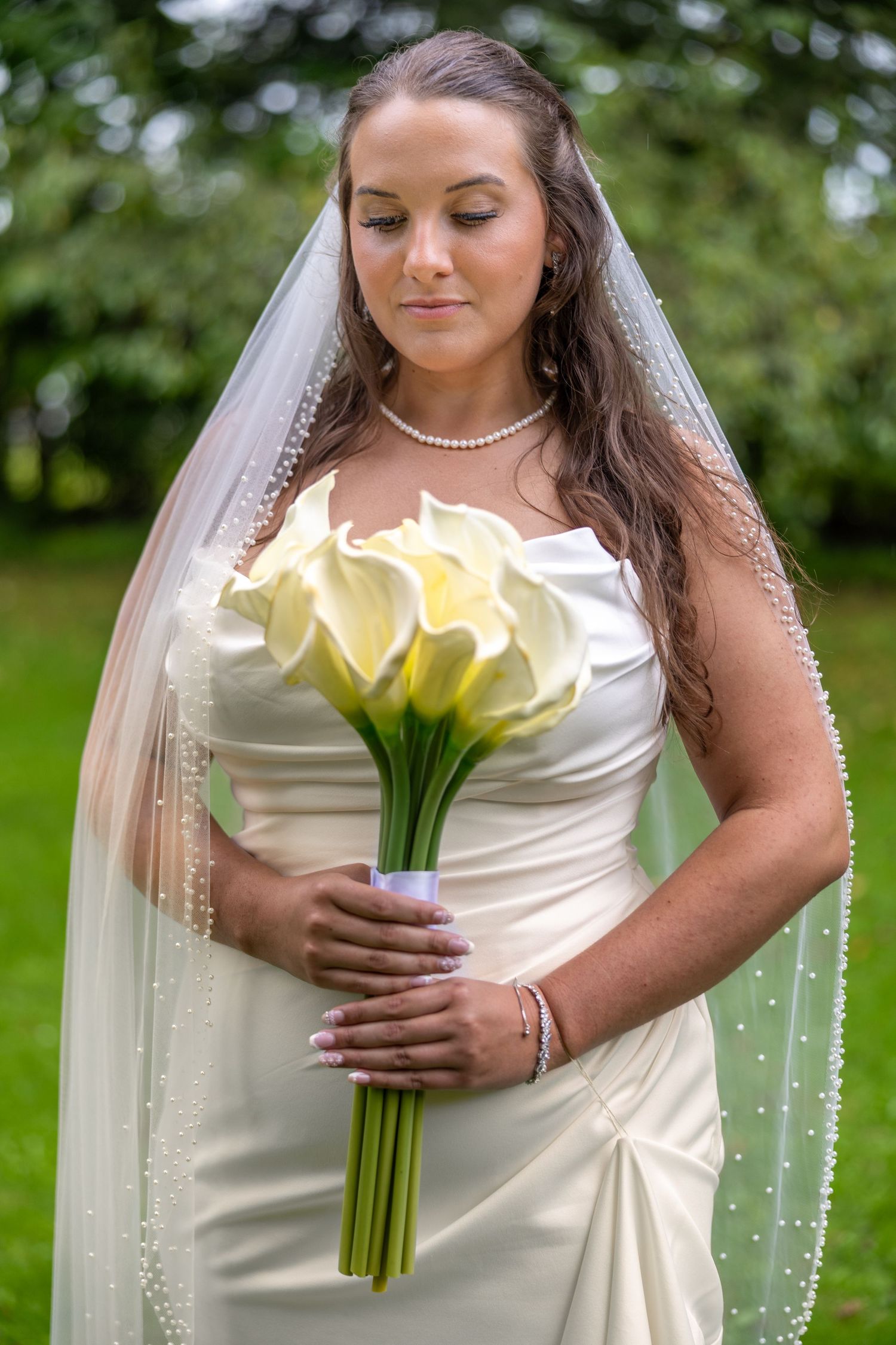 Bride in white satin gown and pearl-edged veil holding calla lily bouquet, standing outdoors in green garden setting.