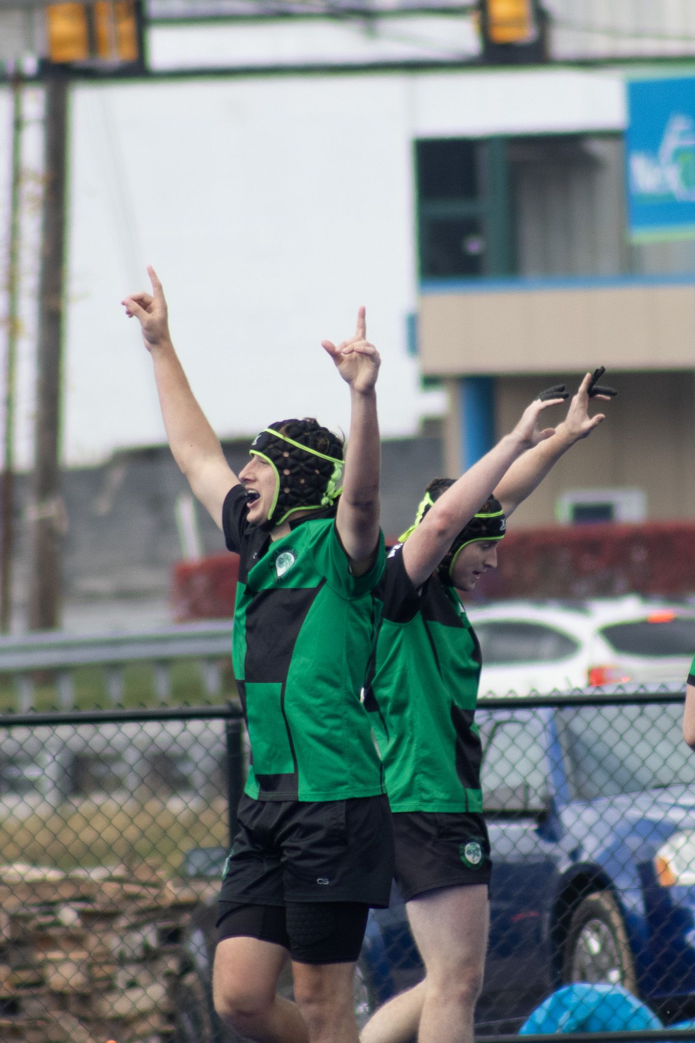 Two youth rugby players in green uniforms celebrate with arms raised during an outdoor match.