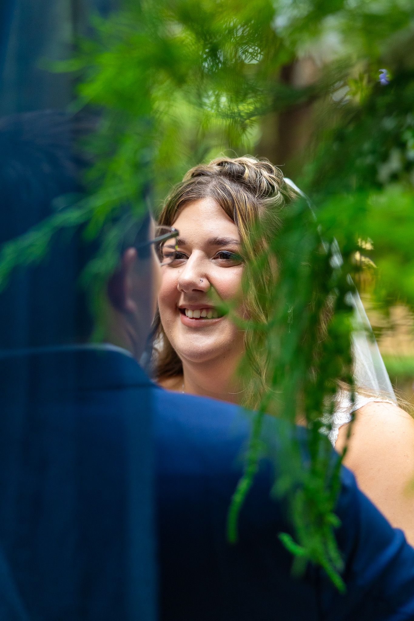 Smiling bride looks at groom through green pine branches during outdoor wedding portrait session.