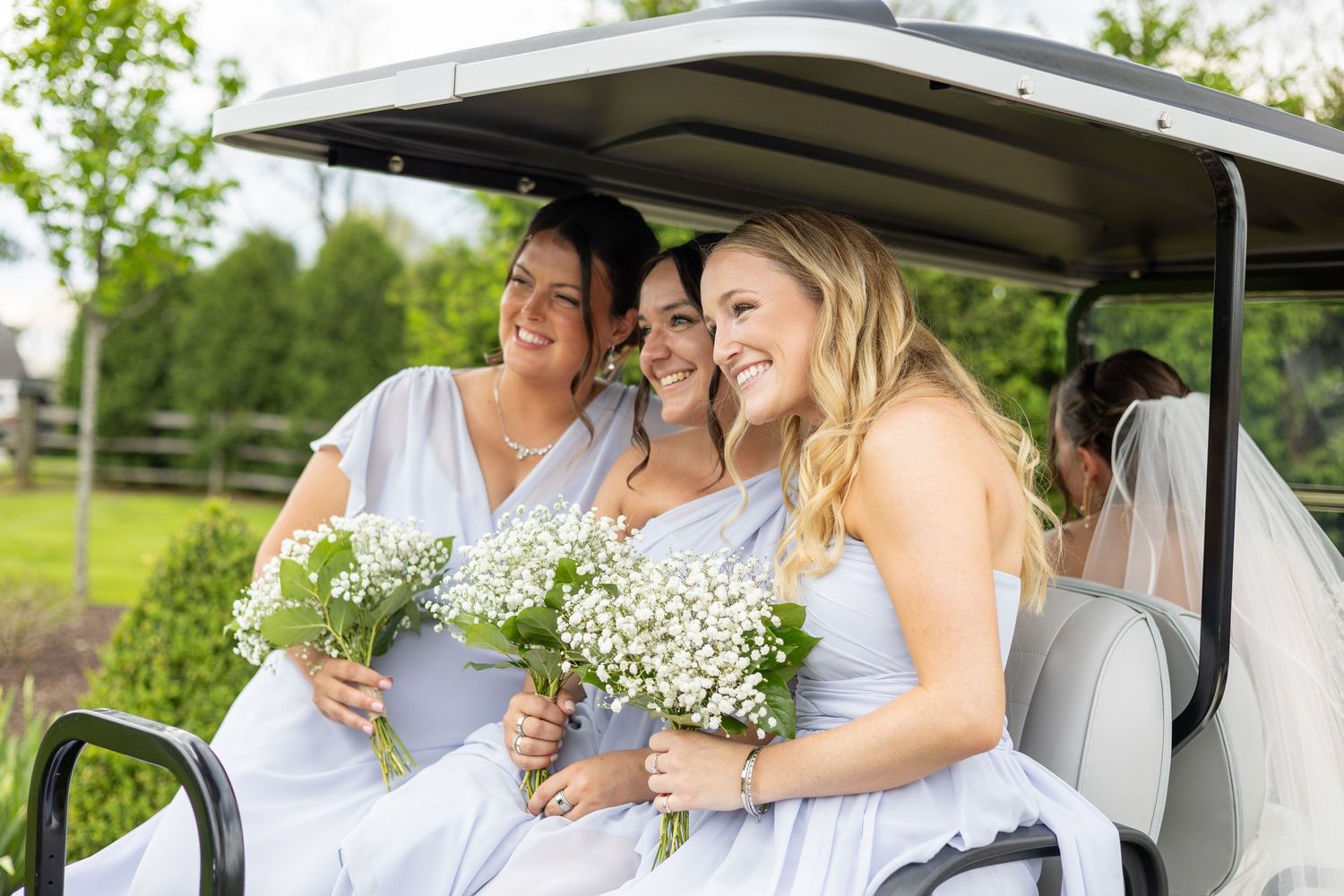 Three bridesmaids in white dresses hold baby's breath bouquets while riding in a golf cart outdoors.