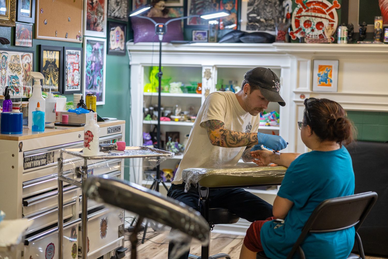 Tattoo artist working on a client's arm in a colorful tattoo shop filled with artwork and supplies.
