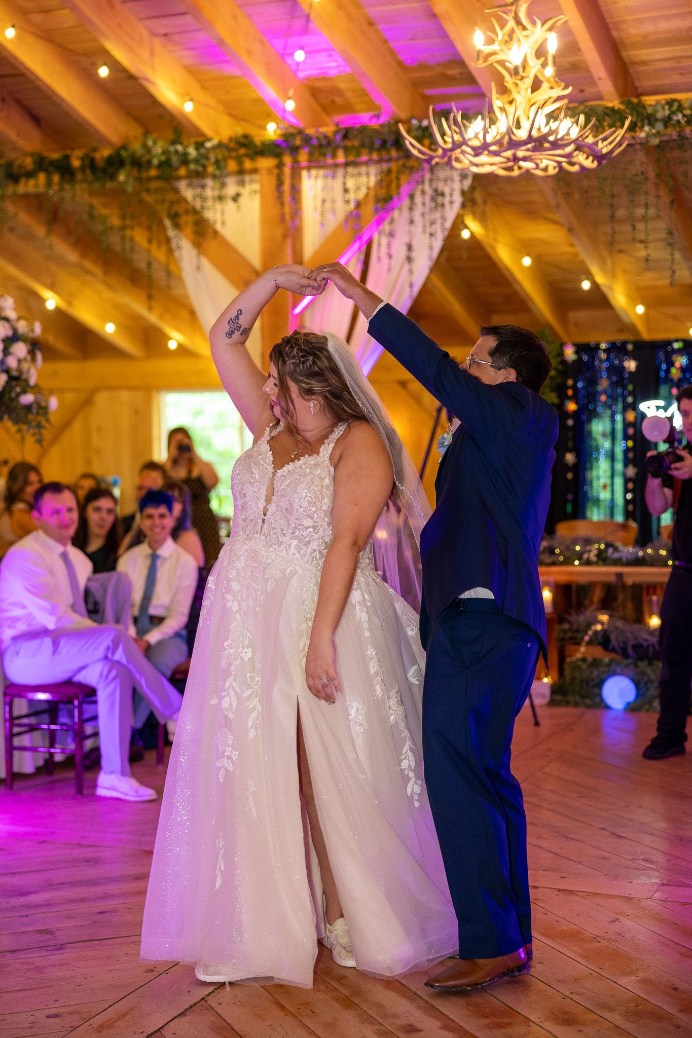 Bride and groom share their first dance in a rustic barn venue adorned with string lights and an antler chandelier.