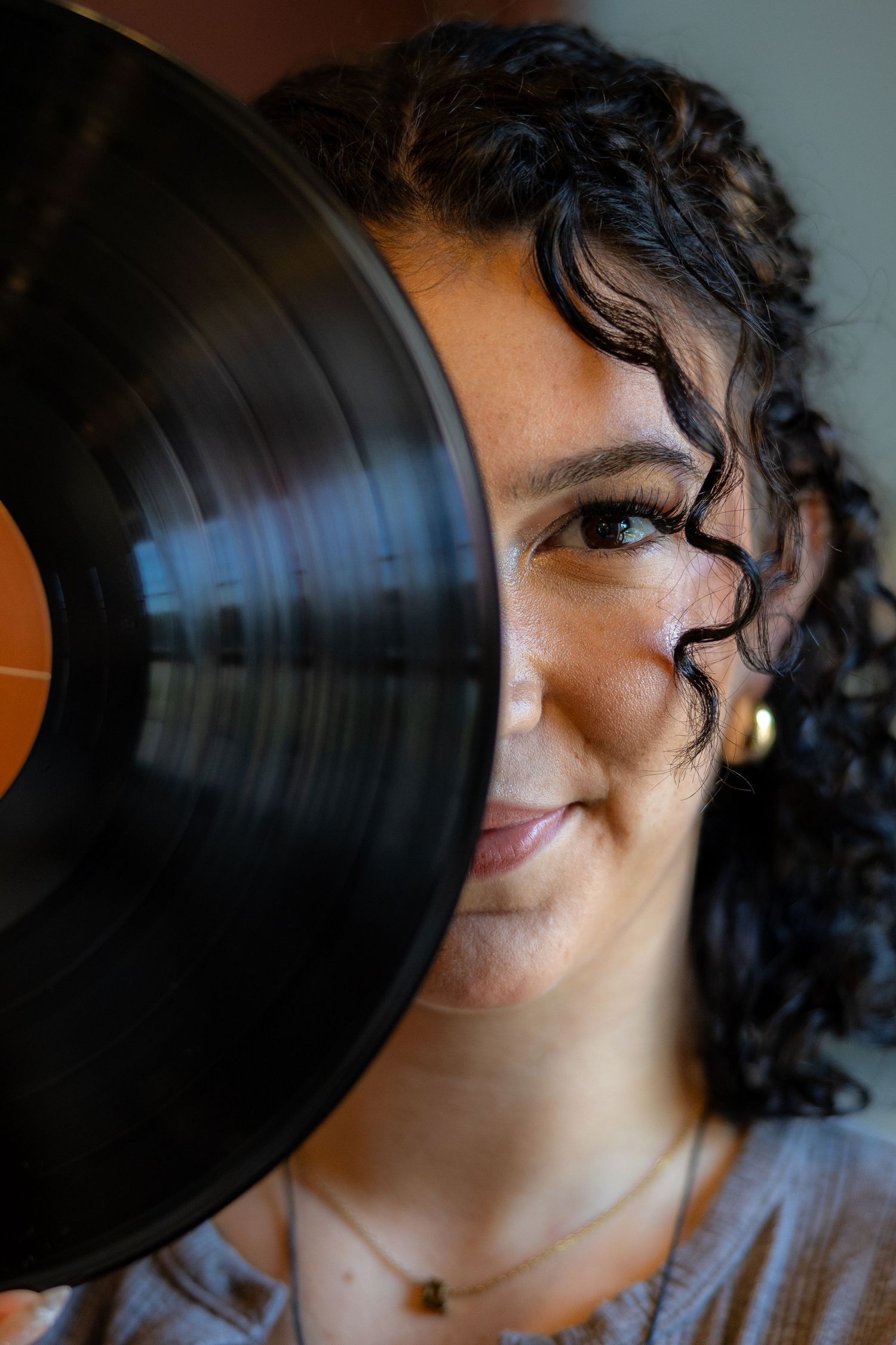 A smiling woman with curly dark hair holds a black vinyl record in front of half her face, wearing a gold earring.